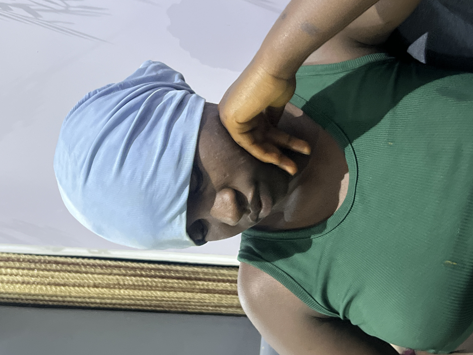 In Gura Top, Nigeria, a young woman with dark skin, wearing a light blue headscarf and a green ribbed tank top, is captured in a close-up, seated portrait. Her head is bowed, with her face turned downwards, and her left hand is raised to her cheek, her fingers gently touching her jawline. The background is a softly lit interior, with a textured, golden-brown curtain on the left and a pale wall with the faint shadow of a plant on the right. The lighting creates subtle shadows and highlights across her face and the fabric of her clothing.