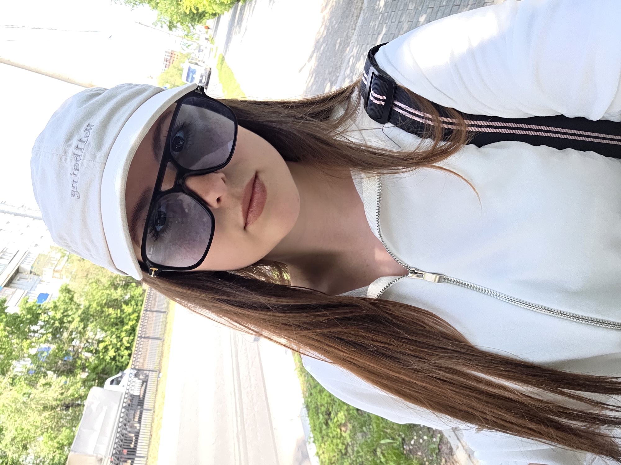 A young woman is pictured in a selfie outdoors. She is wearing a white baseball cap with gray lettering, large black-rimmed sunglasses, and a white zipped jacket. Her long, brown hair falls over her shoulders. A black bag strap with pink stripes is visible on her right shoulder. The background shows a sidewalk, a street with a truck and a car, trees, and buildings. The lighting suggests it is daytime, likely sunny.