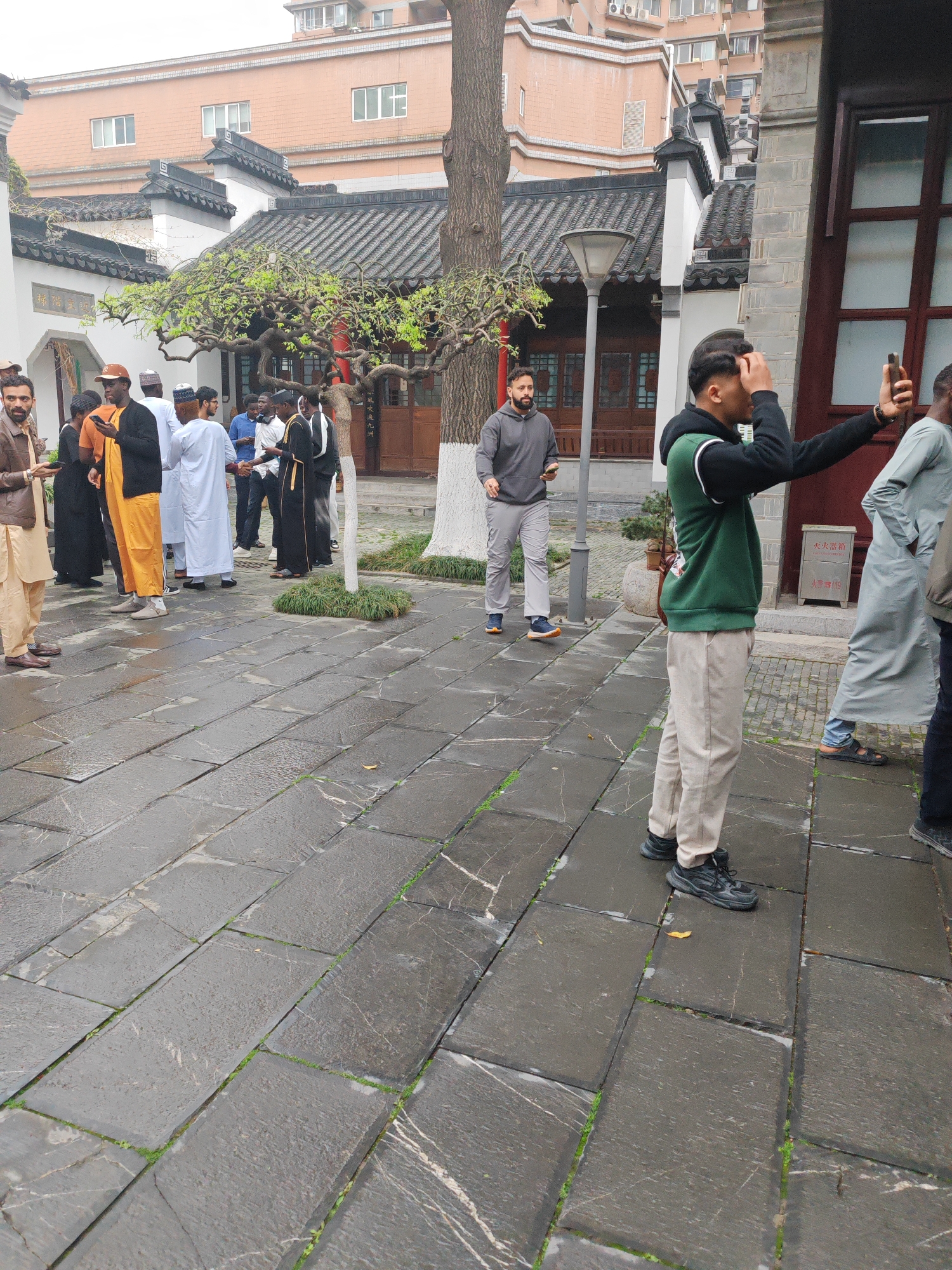 The photograph captures a scene within the city of Qinhuai District, China, likely in a courtyard or open area associated with traditional Chinese architecture. The ground is paved with large, dark stone tiles showing wear and small vegetation growing between the joints.
A group of men, predominantly of African descent, are gathered on the left side of the frame. They are dressed in a variety of traditional Muslim attire, including robes of various colors (white, orange, black), some with head coverings, suggesting a possible cultural or religious gathering. Some are interacting with their phones.
On the right, a younger man wearing a green and black hooded vest over a light-colored long-sleeved shirt, and khaki pants is taking a selfie with his smartphone. Another man, wearing a long grey garment, is partially visible next to him. In the background, a man in a grey sweatsuit walks towards the group.
The architectural backdrop includes traditional Chinese buildings with dark tiled roofs and white walls. A bare tree is also visible.