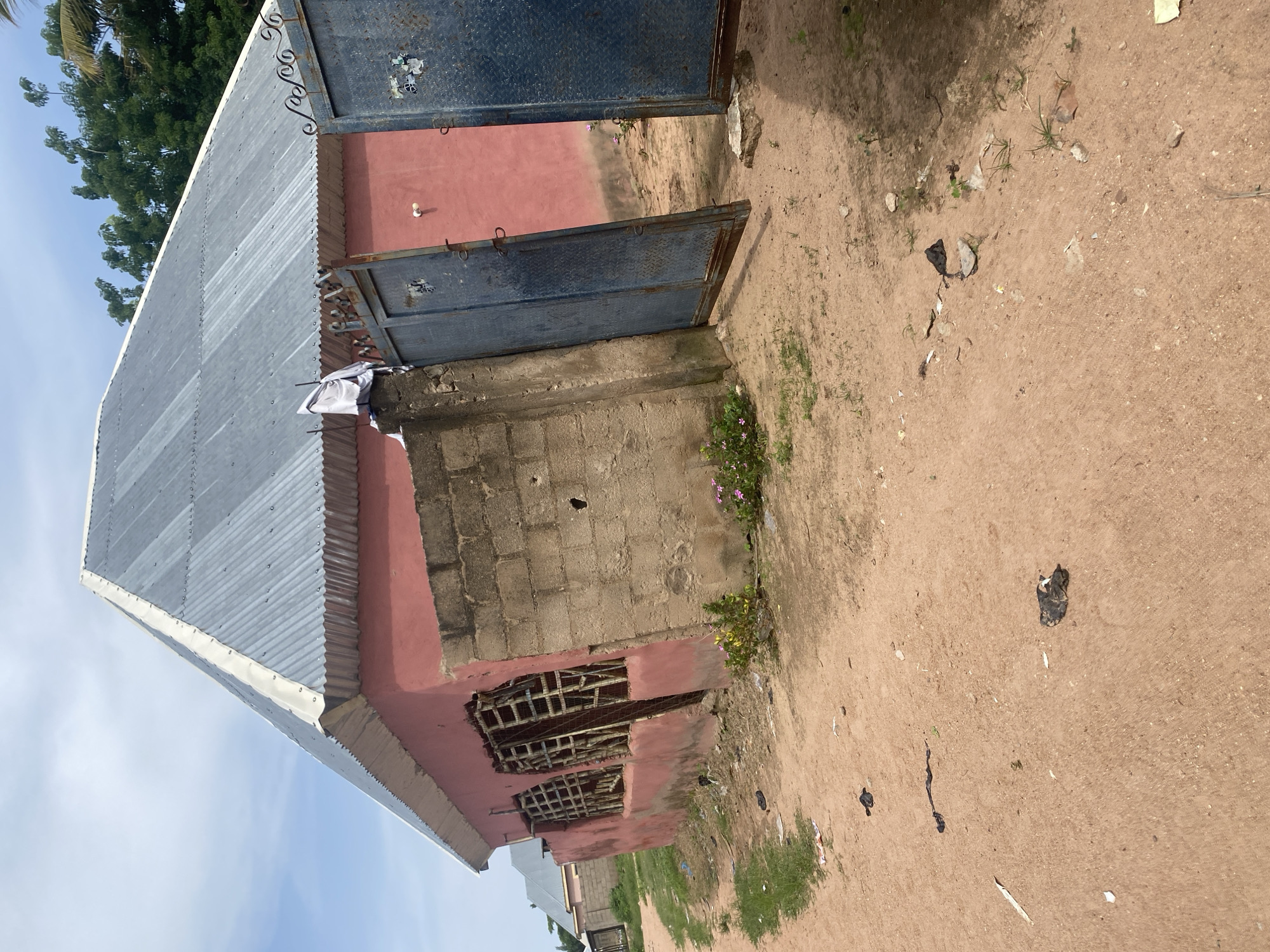 The image depicts a modest building with a corrugated metal roof and pink-painted walls. The building appears to be constructed from concrete blocks in some areas. There are two large, blue metal gates, one of which is open, revealing a further pink-walled structure behind it. Small pink flowers are growing near the building. The ground is sandy and somewhat littered with debris, including pieces of paper and what appears to be a black plastic bag. In the background, there are trees and a clear blue sky, suggesting it is daytime with fair weather. The scene has a rural or semi-rural feel, consistent with the provided location context of Jauro Yinu, Nigeria. No people are visible in the image. The overall impression is one of a simple, functional dwelling or establishment in a developing area.
