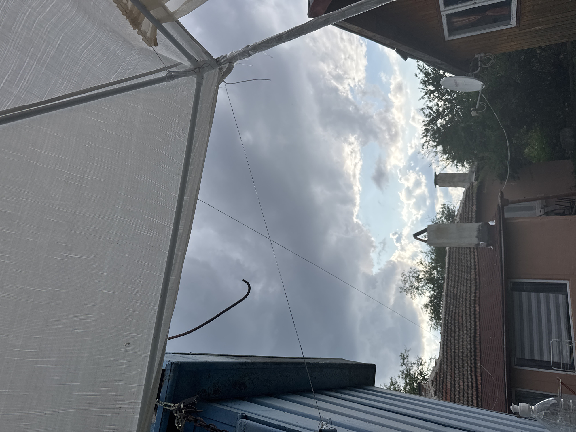 The image depicts a view from under a white tarp, looking towards a cloudy sky with patches of blue. In the foreground, a blue, corrugated metal structure is visible on the left, with a chain attached to it. A bent metal hook hangs from the structure. A thin wire stretches across the frame. In the middle ground, several houses with red tile roofs are visible. One house has two chimneys, and another has a satellite dish mounted on its side. Trees surround the houses. On the far right, a wooden house with a window is visible. The scene appears to be in a residential area in the city of Sredets, Bulgaria.
