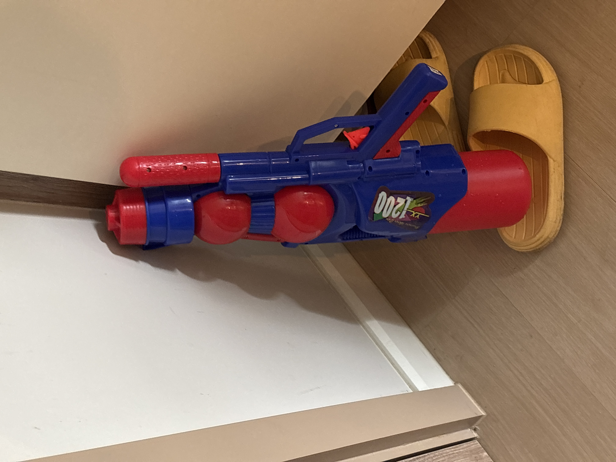 A blue and red water gun rests on a white surface, possibly a shelf or drawer, in what appears to be an indoor environment. Next to the gun are two yellow sandals. The location, Choeng Thale, Thailand, is inferred and not directly evident in the image. There are no people visible in the scene.