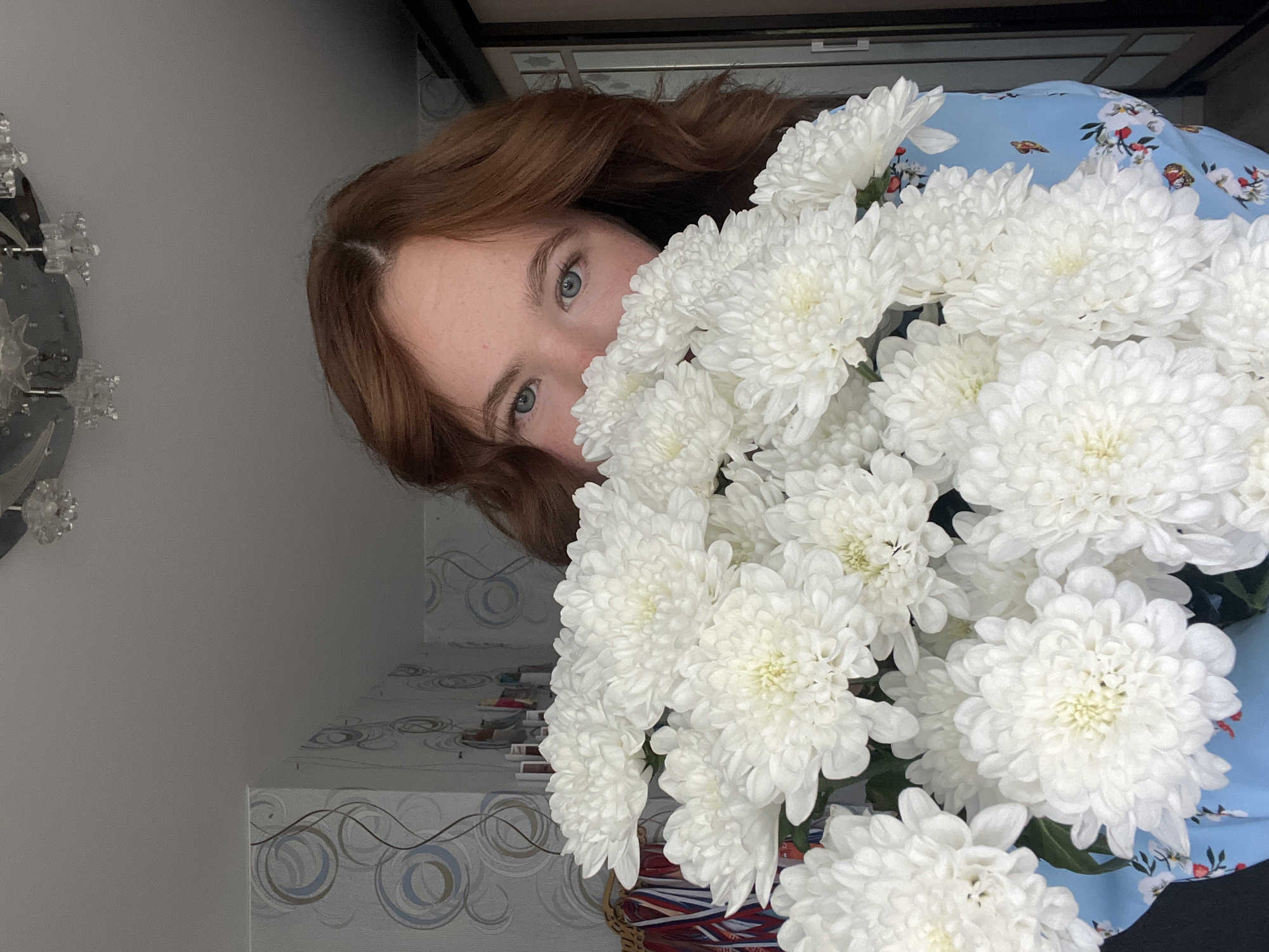 A young woman with auburn hair and blue eyes is holding a large bouquet of white chrysanthemums in front of her face. She is wearing a blue dress with a floral and butterfly pattern. The background shows a room with patterned wallpaper and a chandelier on the ceiling. There is a wardrobe on the right side of the image. The scene appears to be indoors, possibly in a residential setting. The location is Chaikovsky, Russia.