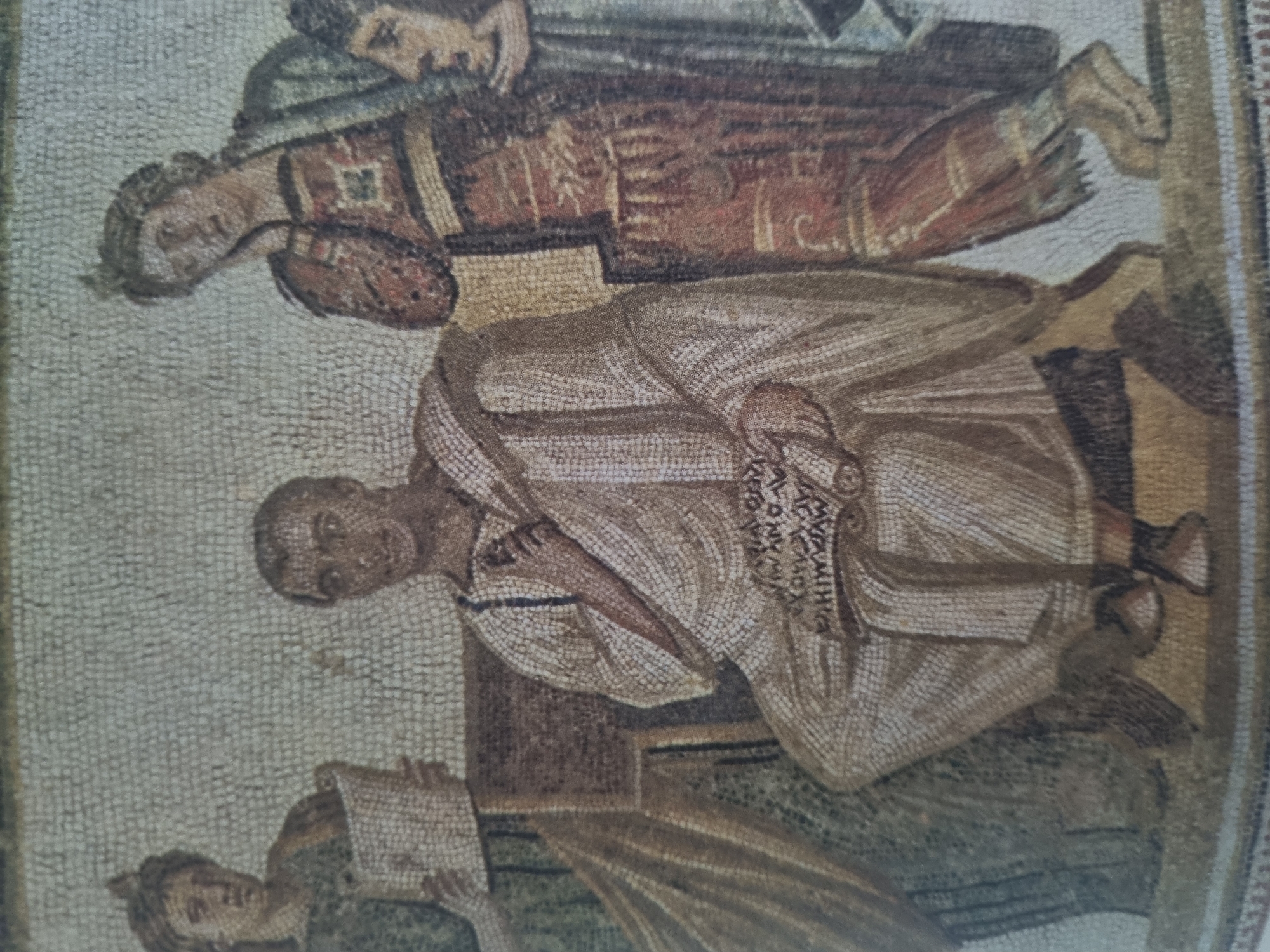 The image is a mosaic depicting several figures. In the center, a man is seated on a chair, wearing a white toga and holding a scroll. To his left, a woman in a green robe stands and holds a similar scroll. To his right, another figure, appearing to be a woman, wears a red and gold patterned garment. The background is a textured mosaic pattern in shades of beige and light gray. The mosaic is framed by a border of smaller tesserae. The scene appears to be an interior setting, possibly a library or study, given the presence of scrolls and the seated figure. The figures are rendered in a stylized, ancient art form, with details conveyed through the arrangement of small stone or glass pieces.
