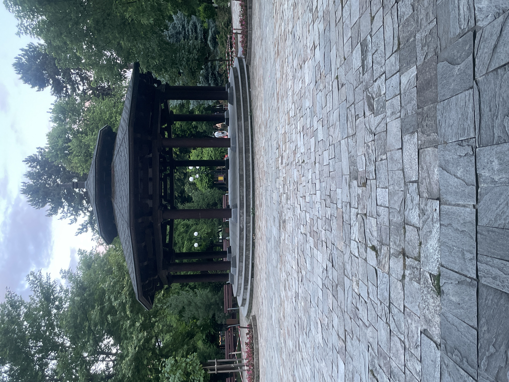 The image depicts a large, octagonal wooden gazebo with a dark, multi-tiered roof supported by thick pillars. The gazebo is situated on a raised, circular platform accessed by several steps. The platform and the area in front of it are paved with irregular, grey flagstones. Surrounding the gazebo are lush green trees of various species, with some evergreen trees also visible. In the background, there are park benches, lampposts, and a glimpse of playground equipment. Two individuals are visible walking near the gazebo. The scene appears to be set in a park, likely during twilight or a cloudy day, given the soft lighting and diffused sky. This is the central gazebo in the city of Gotse Delchev, Bulgaria, a common gathering place for residents.