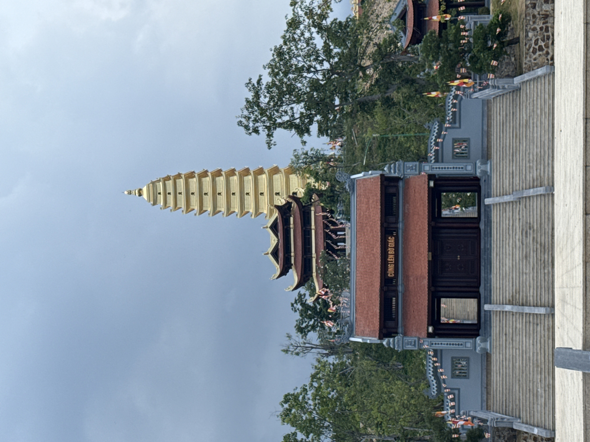 The image shows a tall, golden pagoda in Nha Trang, Vietnam. The pagoda has multiple tiers and tapers to a point. Adjacent to the pagoda is a structure with a red-tiled roof and dark wood doors. Stairs lead up to this structure. Trees surround the buildings, and colorful banners are strung around the area. The sky is blue with some clouds. The gate reads, "Cung len Bo Giac".