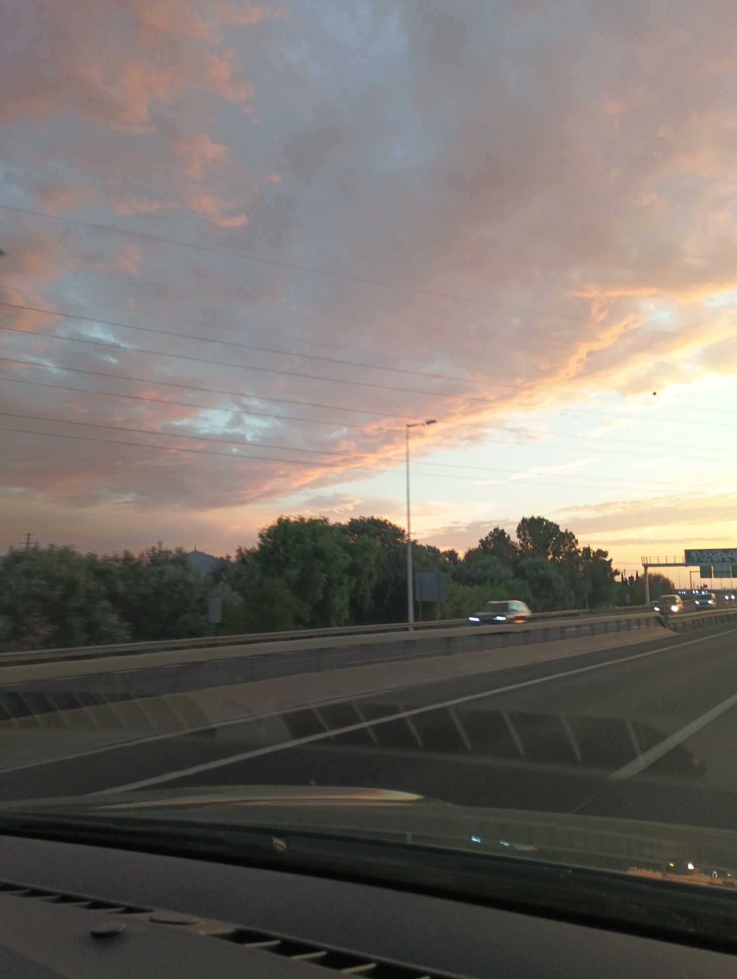 The image depicts a highway scene at dusk. The sky is filled with a mix of soft pink, orange, and blue clouds, suggesting the sun is setting. Several power lines are visible across the upper portion of the sky. On the left side of the highway, there is a dense line of green trees, with a tall lamppost positioned near the road. On the right side of the highway, more trees are visible, along with highway signs and streetlights, some of which are illuminated. Several cars are driving on the highway, their headlights visible. The foreground shows the interior of a vehicle, likely a car, with a dashboard visible. The overall impression is a view from a moving vehicle on a highway during sunset.