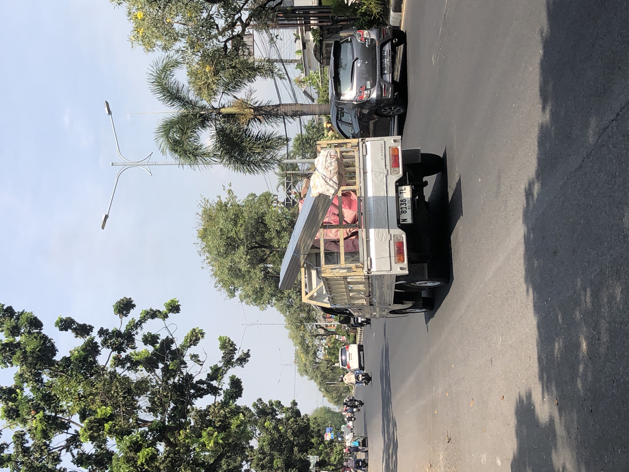 The image depicts a street scene in Larangan, Indonesia. A white pickup truck with wooden sides is driving on the right side of a paved road. The truck is carrying a load of what appears to be construction materials, including long metal beams and several large, wrapped pink items. The truck's license plate reads "N 8138 TL". To the right of the truck, a dark gray hatchback car is also traveling in the same direction. The car's license plate is partially visible as "W 1705 XZ".

The road is lined with trees on both sides, casting shadows on the asphalt. In the background, palm trees are visible, along with streetlights and utility poles with many wires. Other vehicles, including motorcycles and cars, are seen on the road further ahead, suggesting moderate traffic. The sky is clear and bright, indicating a sunny day. The environment appears to be an urban or suburban area with residential buildings and some commercial structures visible in the distance.