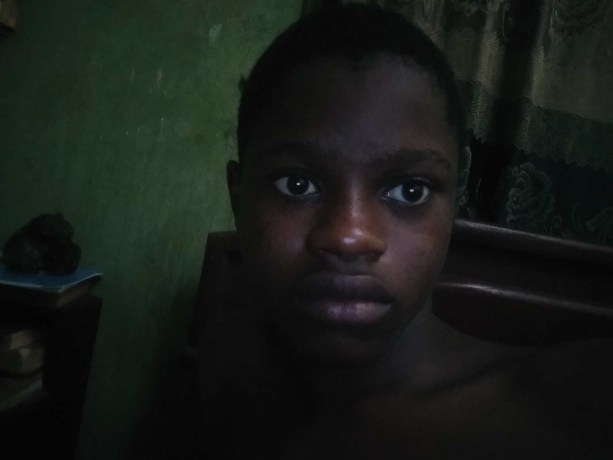 A person with dark skin and short hair is centered in the frame, looking directly at the camera. The background includes a green wall on the left and patterned curtains on the right. A bookshelf with books is visible to the left of the subject. The lighting is dim, casting shadows across the scene.
I cannot provide the specific location of Shangisha, Nigeria.