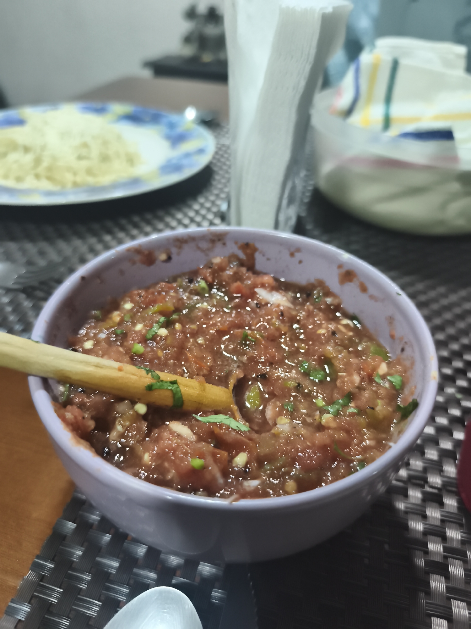 The image depicts a bowl of salsa with a wooden spoon, positioned prominently on a table. The salsa appears to be a fresh, homemade variety, containing diced tomatoes, onions, and green herbs, likely cilantro. In the background, a plate of what seems to be cooked noodles is visible, along with a container of napkins and a covered dish. The setting is indoors, possibly a dining room or kitchen in Mexico City, Mexico. The lighting is soft and diffused, suggesting daytime. The scene focuses on the food arrangement, suggesting a meal is being prepared or served.