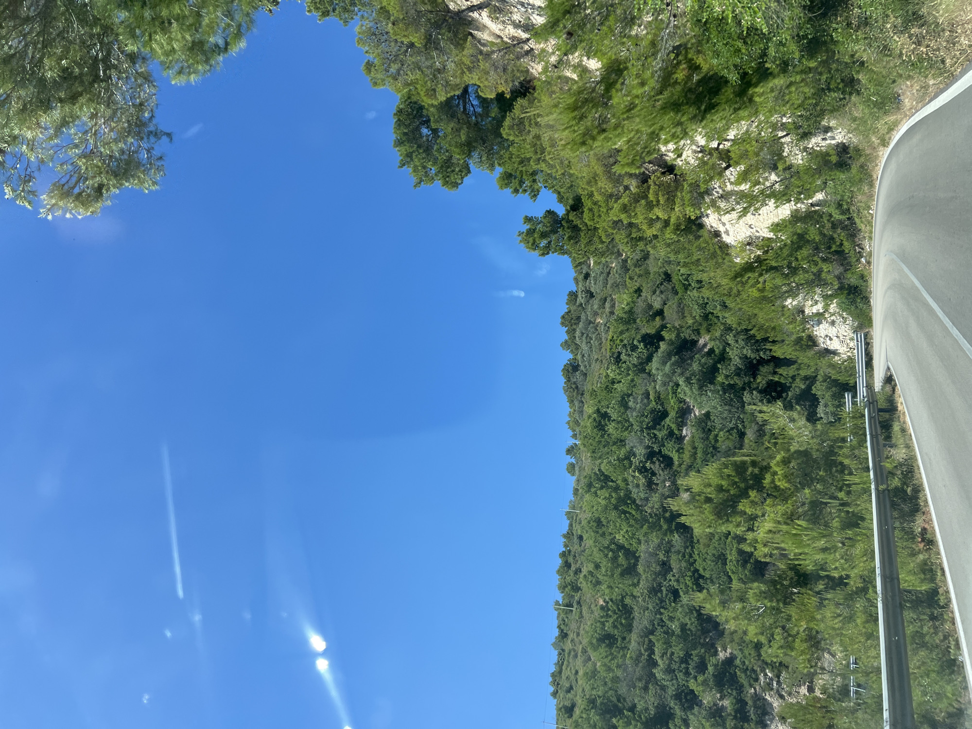 This image captures a scenic view of a curved road passing through a lush, green hillside under a clear blue sky. The road is paved and marked with white lines, suggesting it's a route for vehicles. A metal guardrail runs along the left side of the road, separating it from the steep incline. The hillside is densely covered with various types of trees and vegetation, with some rocky outcrops visible. The sky is a vibrant blue, with a few lens flares appearing as streaks of light. The presence of bright sunlight indicates a daytime scene. The vegetation and terrain are consistent with the Mediterranean landscape found in areas like Mattinata, Italy, known for its rugged coastline and verdant hills. There are no people or animals visible in the image.