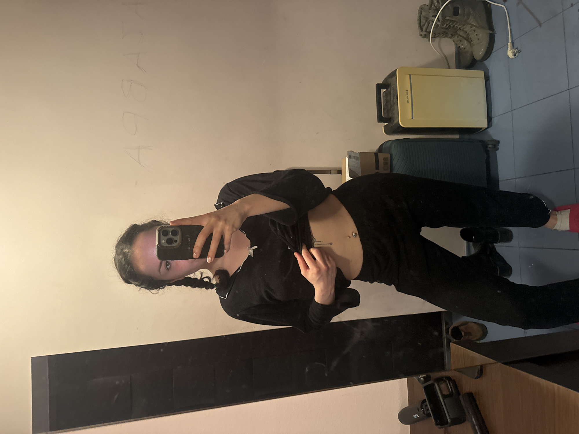 A young woman with pigtails is taking a selfie in a mirror. She is wearing black clothing and has a belly button piercing. The room appears to be a messy interior with luggage, shoes, and an electronic device in the background. There is writing on the wall in the upper left corner. The location cannot be determined.