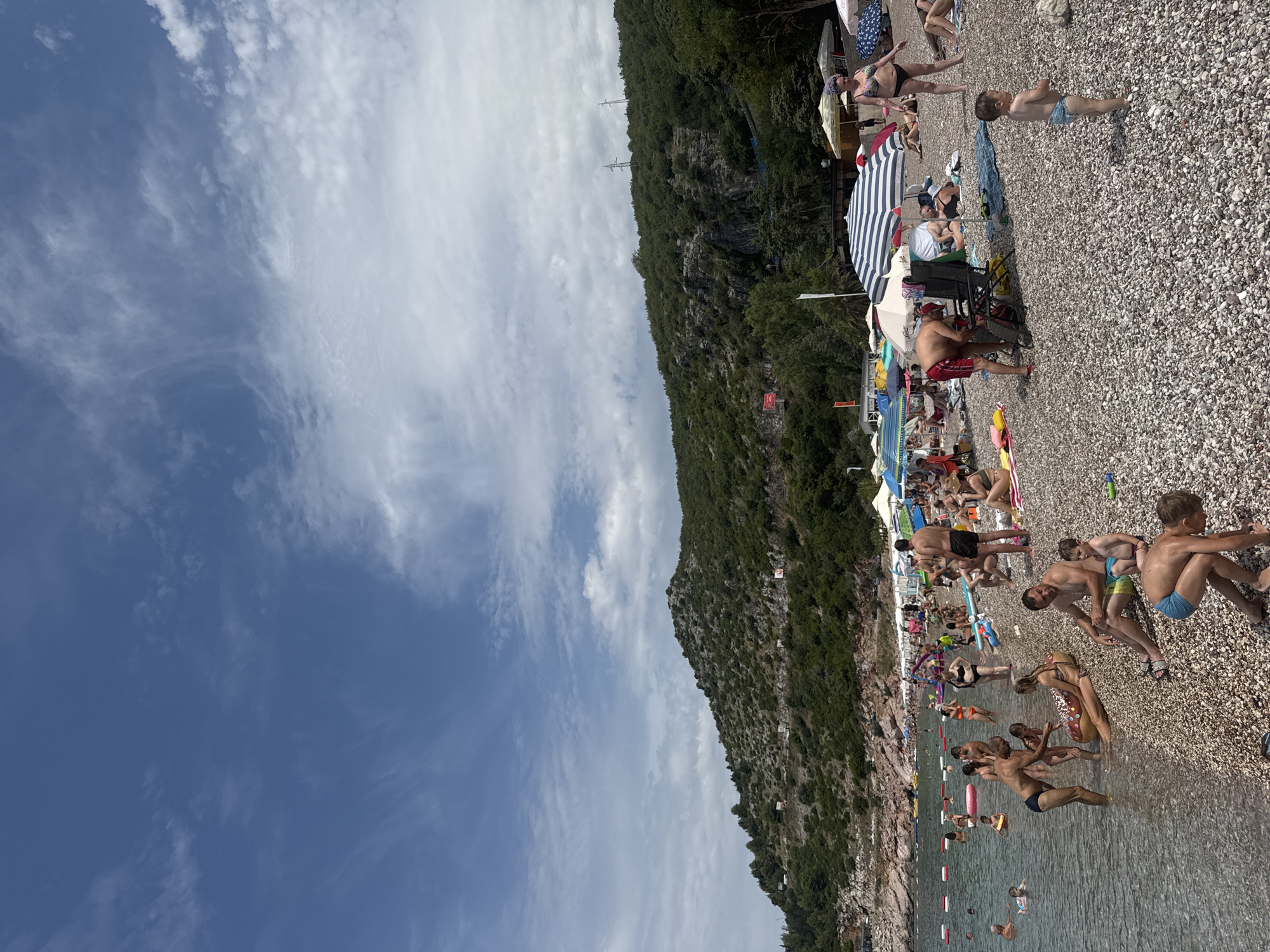 This image depicts a lively beach scene in Kamenovo, Montenegro. The beach is covered in small, grey pebbles, and the water is clear and calm. Numerous people are present, enjoying the sunny weather.
In the foreground, several individuals are on the beach, some sitting under umbrellas, others wading in the shallow water. A man in red shorts is tending to a beach chair, while a young child in blue swim trunks plays on the pebbles. To the right, a woman in a black bikini stands near an umbrella.
Further back, the beach is more densely populated. People are spread out on towels and beach chairs, with many in the water, swimming or playing. Inflatable toys, including a large donut-shaped float, are visible. Beach umbrellas of various colors and designs provide shade.
The background features a rocky, tree-covered mountainside that slopes down towards the sea. The vegetation on the hillside is dense and green. A road can be seen winding along the mountainside, with some small buildings and structures visible in the distance. The sky is blue with scattered white clouds.