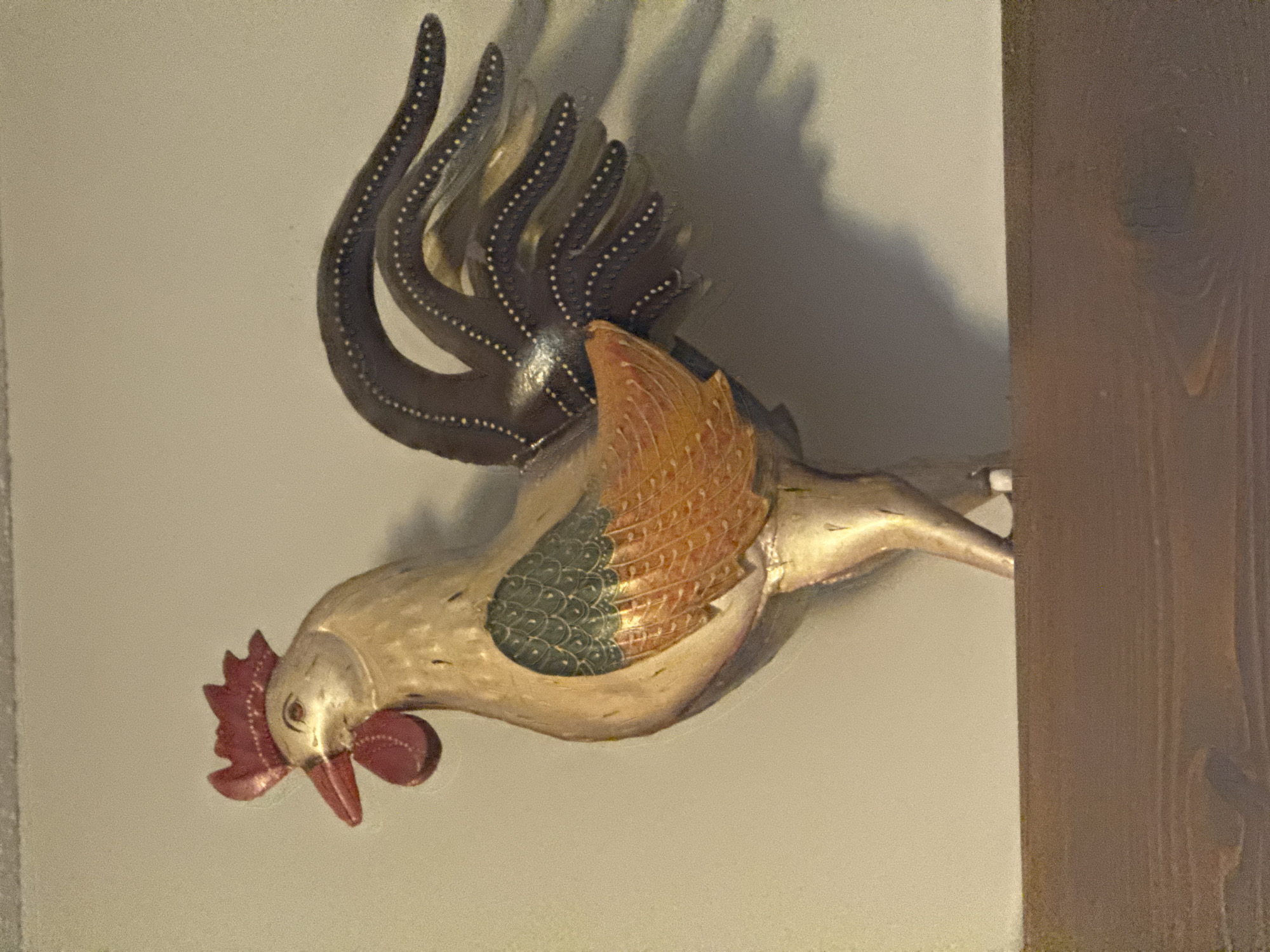 A metallic rooster ornament, predominantly gold, stands on a dark brown wooden shelf against a light grey wall. The rooster has a red comb and wattle, a red beak, and its wings are detailed with green and orange feathers. Its tail is composed of dark, curved metallic feathers, each dotted with white. The ornament casts a shadow on the wall behind it. The image does not provide any information about the location within the Town of Cochrane, Canada.