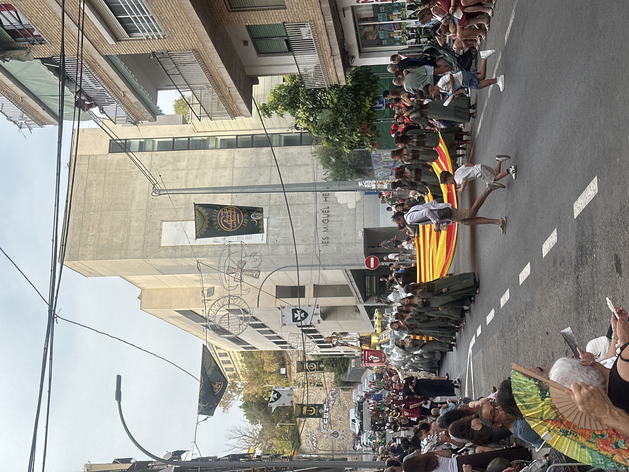 The image depicts a street procession in Alicante, Spain, likely a festival or parade. In the background, a large, modern concrete building with the words "IES MIGUEL HERNÁNDEZ" is visible, indicating the location as a public institution, possibly a school. The street is lined with spectators seated on chairs and standing. A procession is moving down the street, featuring people in historical or traditional costumes, including women in long, dark green dresses and men in maroon garments. A large yellow and red striped flag, possibly the Senyera (flag of Catalonia or Valencia), is being carried by several people. Two young boys are running alongside the flag. In the foreground, spectators are watching the procession, with one person holding an open fan. The scene is outdoors, with overhead wires and a lamppost visible.