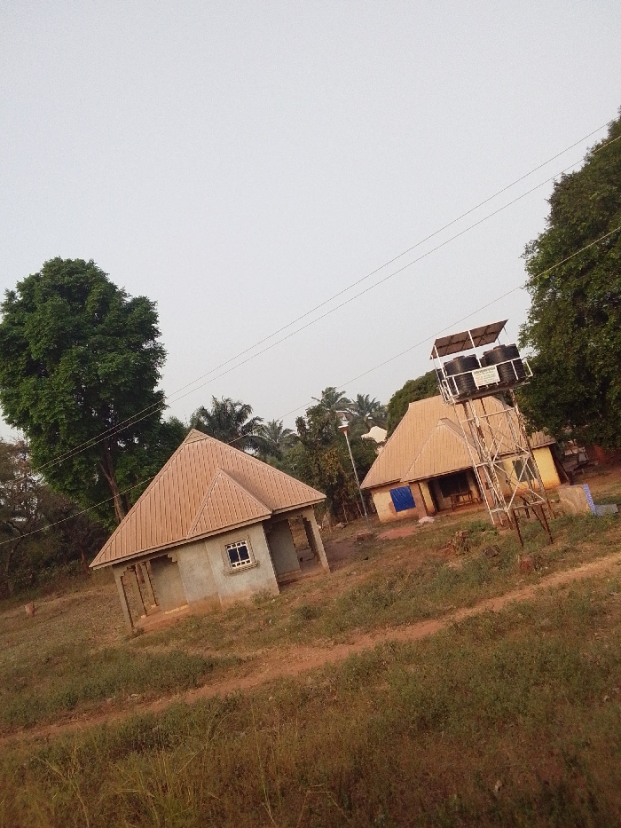 This image depicts a semi-rural landscape in Ankpa, Nigeria, under an overcast, light grey sky. The ground consists of reddish-brown soil with scattered patches of dry and green low-lying vegetation.
Two single-story structures with light brown corrugated metal hip roofs are visible. The building on the left features unpainted grey concrete block walls, a single rectangular window with white framing, and an open front porch supported by pillars. To its right, another building with a similar roof appears; a blue tarp or fabric covers a portion of its exterior, possibly an entrance or window.
Between these two buildings, a tall metal lattice tower stands, supporting two black cylindrical water tanks and a framework of several dark rectangular solar panels beneath a light-colored canopy, indicative of a solar-powered water system. A green sign with white text is affixed to the tower's support structure. A tall street light pole with a luminaire is positioned near the second building.
Overhead, three thin electrical wires run horizontally across the frame. Large, leafy green trees, including discernible palm trees, are present on the left and right edges, as well as in the background. No individuals are present in the scene.