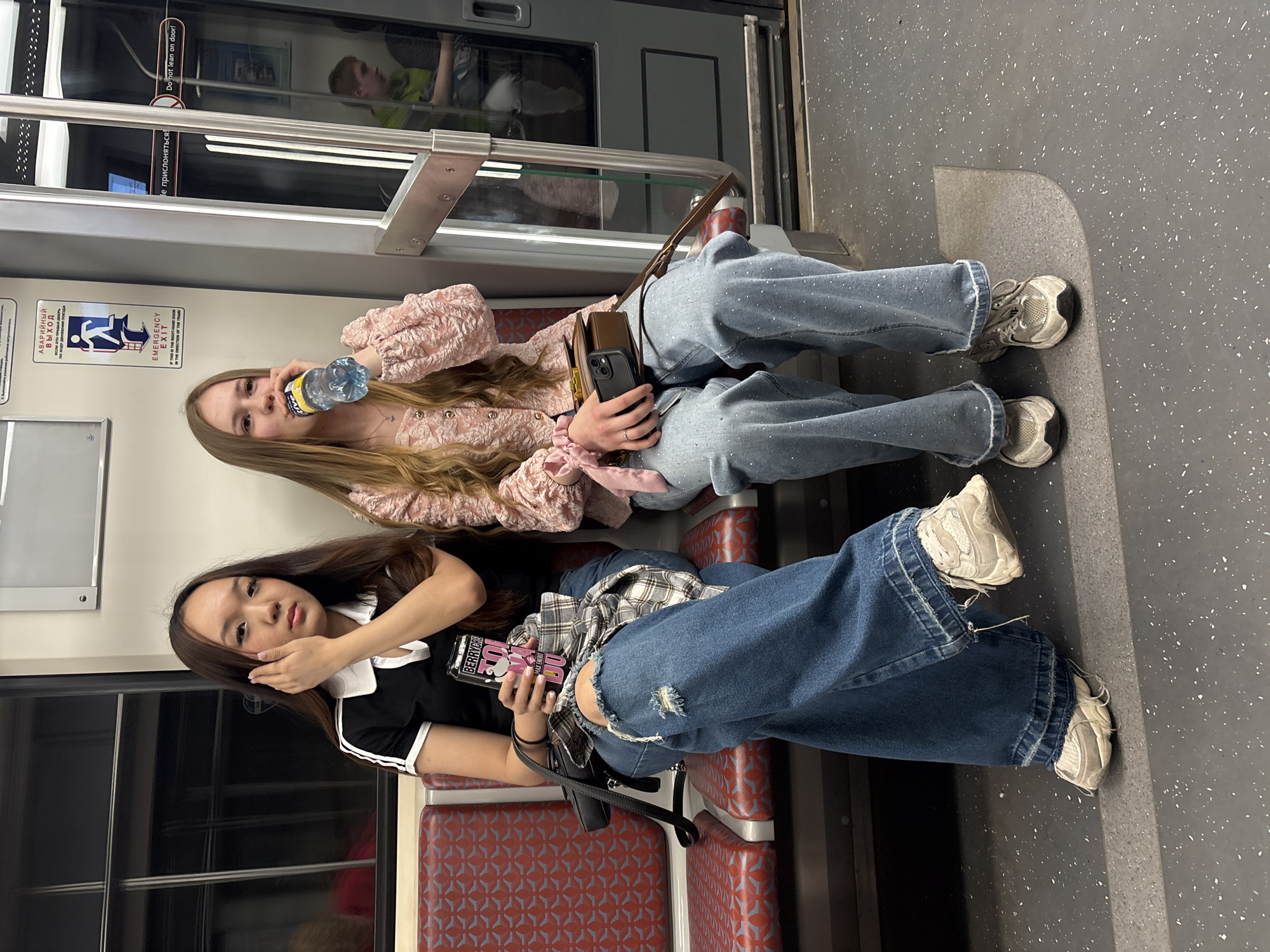 Two young women are seated on a red upholstered bench with a grey geometric pattern, inside a metro car in Saint Petersburg, Russia. The car features white upper wall panels, dark grey lower panels, and dark-tinted windows framed in black. An "EMERGENCY EXIT" sign, written in both Russian ("АВАРИЙНЫЙ ВЫХОД") and English, is affixed to the wall above the right subject, alongside a "Do not lean on door!" sign also in Russian ("Не прислоняться!") and English.
The subject on the left, identified as East Asian or Southeast Asian, has long dark brown hair with lighter streaks, parted in the middle. She wears a black short-sleeved polo shirt with a white collar and two white stripes on the sleeves, paired with blue wide-leg ripped jeans. Her left hand holds a pink can with "BERRYGRA" and "MAX ENERGY" visible on the label, while her right hand is raised, touching her right cheek. A black shoulder bag with a metal buckle strap rests on her lap, and a plaid shirt in shades of grey, black, and pink is draped over her jeans. She wears white chunky sneakers with grey accents.
The subject on the right, identified as Caucasian, has long wavy blonde hair. She wears a pink textured long-sleeved top with puff sleeves and gold buttons, and light blue wide-leg jeans embellished with small white studs or rhinestones. She is actively drinking from a clear plastic water bottle with a blue cap and a yellow label (partially visible "TYMA"). A small brown leather shoulder bag with a gold clasp sits on her lap, topped by a black smartphone in a brown case. A pink scrunchie is on her left wrist, and she wears grey chunky sneakers.
In the reflection of the train door window to the right of the women, a young male with light hair, wearing a lime green top, is visible seated further down the car. The floor of the metro car is dark grey with white speckles, and a light grey rectangular outline is present on the floor near the right subject's feet.