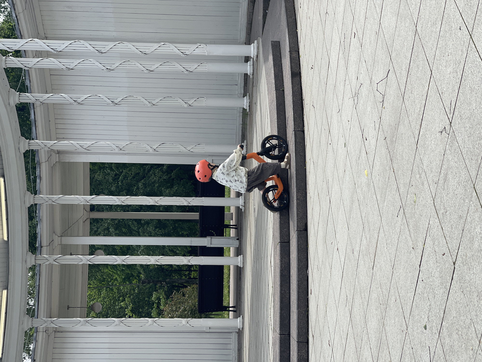A full shot captures a young person on an orange balance bike, positioned near a white colonnade structure, with a grey stone-tiled ground stretching into the distance. The person is wearing an orange helmet, patterned shirt, and striped pants. They are situated on the right side of the frame, near a set of steps that lead to the colonnade. The colonnade has white columns with decorative horizontal bands and a white paneled wall. Benches are visible behind the colonnade. The background features green trees, and the scene is bathed in sunlight.