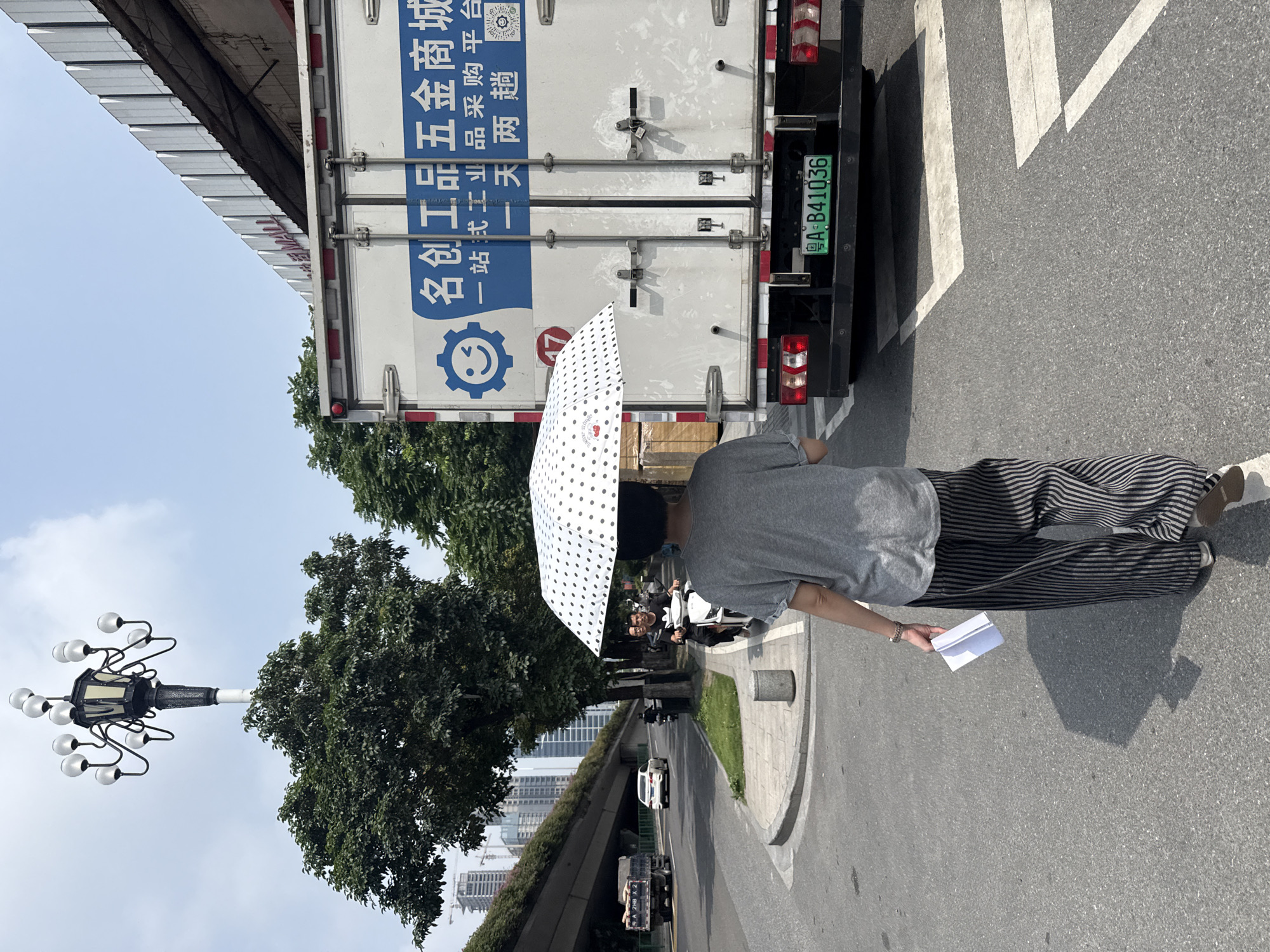 In a city scene in Tianhe District, China, a person walks away from the camera under a white umbrella with black polka dots, holding a folded piece of white paper in their left hand. They are wearing a gray t-shirt and black and white striped wide-leg pants. The person is walking on a crosswalk in front of a large white truck. The truck has blue Chinese characters and a blue logo on its side. The license plate of the truck is green and reads "A B41036". Behind the truck, a white building with a corrugated roof is visible. To the left of the truck, trees line the sidewalk and street. In the background, a highway overpass with cars can be seen, and tall buildings are visible in the distance. A traditional lamppost with multiple lights stands above the trees. The sky is clear and blue.