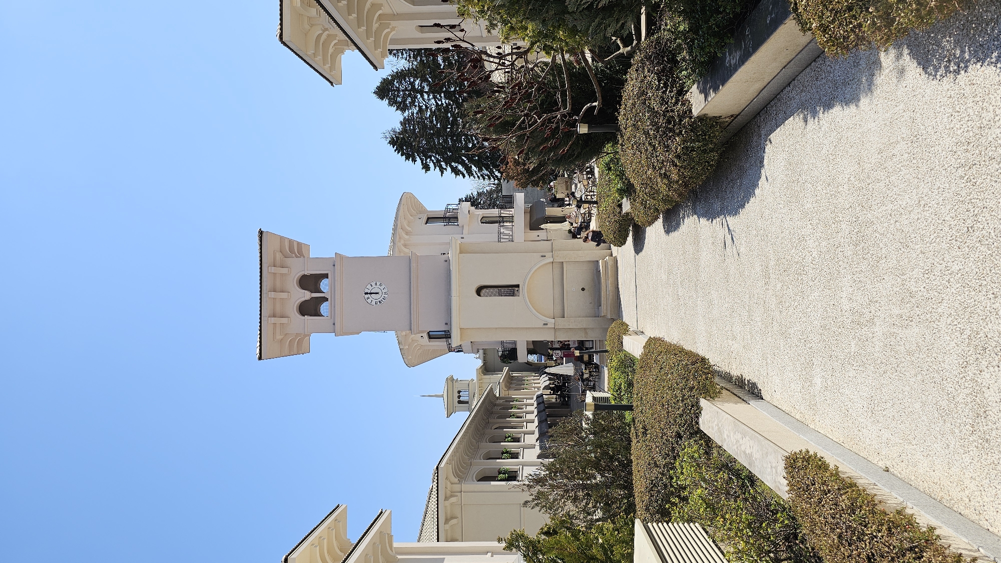 The image shows an outdoor view of the city of Varna, Bulgaria. The focus is on a light-colored building with a clock tower. The tower is tall and slender, with a clock face visible near the top. The building is surrounded by trees and greenery, and there is a walking path that leads towards the building. People are visible at tables, suggesting a cafe or restaurant setting. The sky is clear and blue.