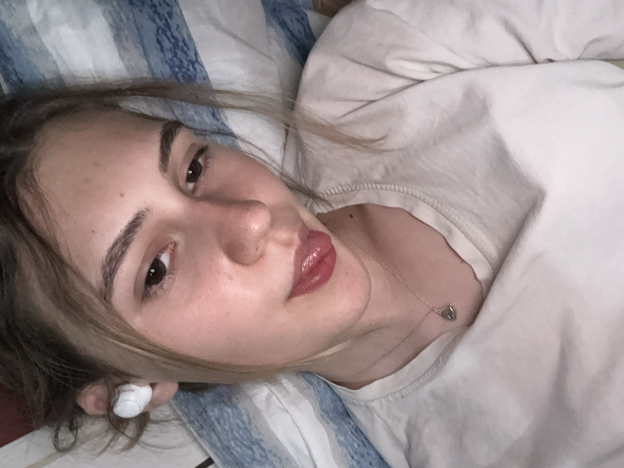 The image shows a close-up, angled selfie of a young woman lying down, with her head slightly tilted and her tongue sticking out. She has light brown hair pulled up loosely, and is wearing white earbuds in her left ear. She is wearing a light-colored t-shirt and a delicate silver necklace with a heart-shaped pendant. The background appears to be a bed with blue and white striped bedding. The overall lighting is soft and appears to be artificial, casting a warm glow on her face. There is no discernible location in the city of Panagyurishte, Bulgaria, or any other specific geographical markers in the image.