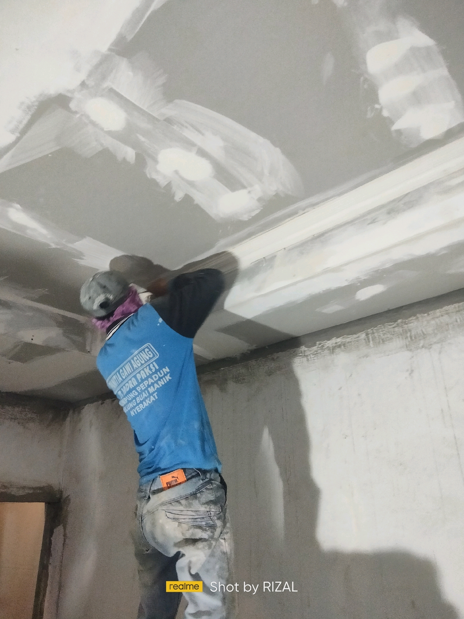 A construction worker, seen from the back, is applying plaster to a white ceiling with a trowel. He is wearing a blue t-shirt, jeans, a grey baseball cap, and a purple scarf around his neck. The ceiling appears to be a drop ceiling with a decorative molding. The walls are bare concrete, and the room is under construction. The image is taken from a low angle, looking up at the ceiling. The bottom right corner of the image has a watermark that reads "realme Shot by RIZAL".