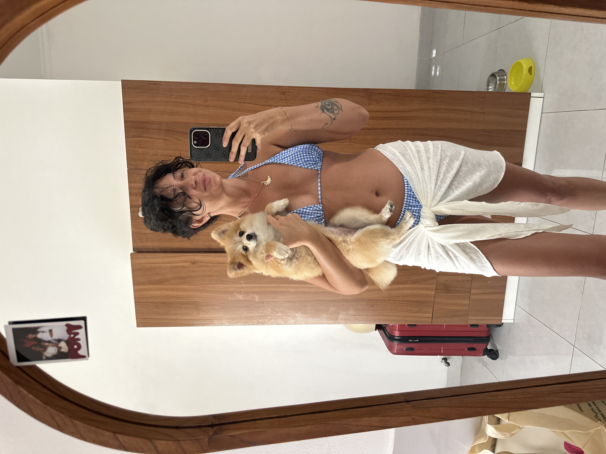 A woman stands in front of a mirror in a room, taking a selfie. She is holding a small, fluffy dog in her arms. The woman is wearing a blue and white checkered bikini and a white sarong tied around her waist. Her hair is dark and curly, and she has a necklace with a sun pendant. In the background, there is a wooden wardrobe, a red suitcase, and a canvas bag. On the wall above the wardrobe, a small framed picture of three people is attached. The floor is tiled. This scene appears to be in a residential setting, possibly a hotel room, in Kuta Selatan, Indonesia, given the presence of a suitcase and the general aesthetic.