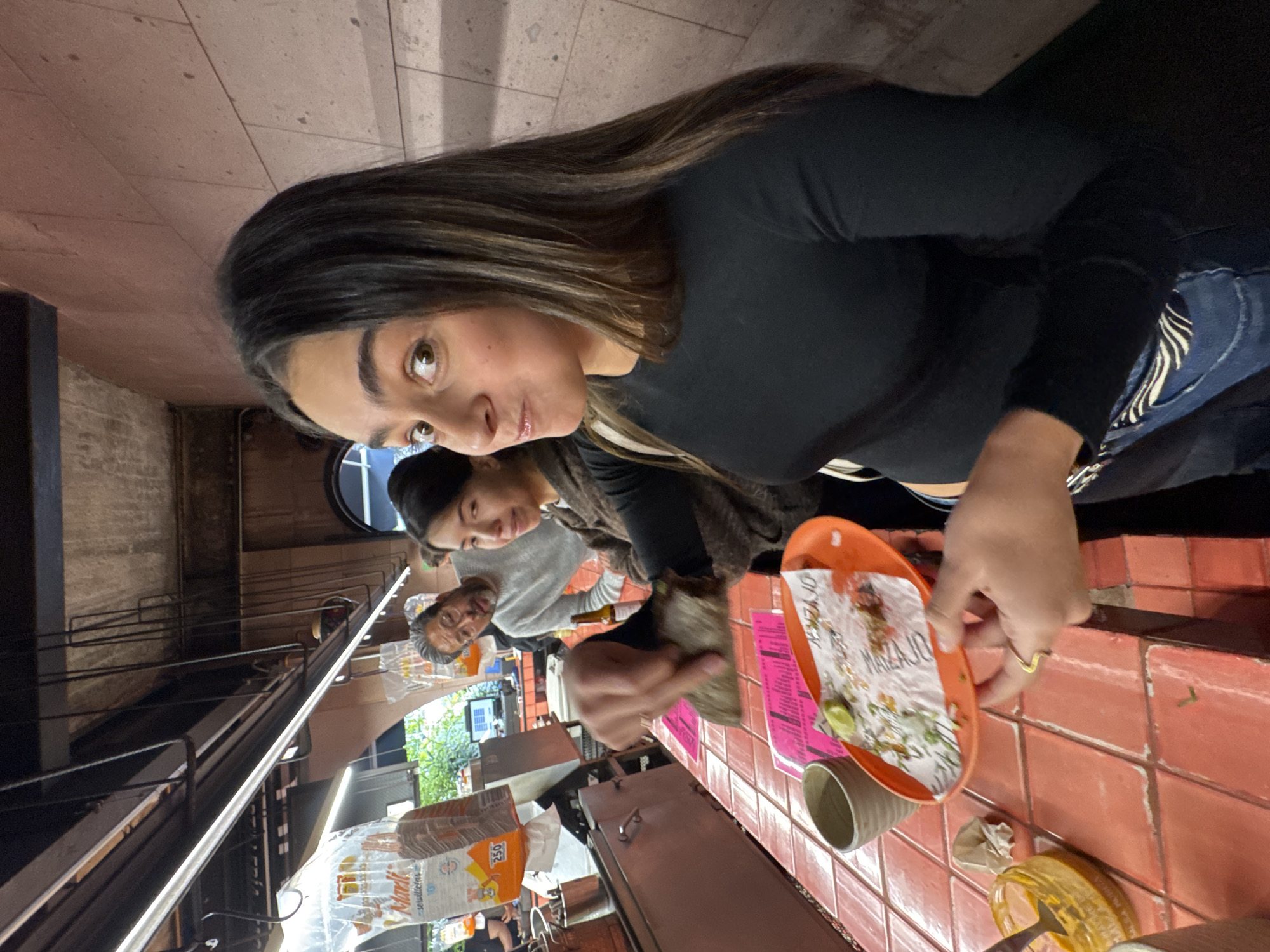 A tilted, close-up indoor shot shows a young woman eating a meal at a restaurant in Mexico City, Mexico. The restaurant appears to be indoors but connected to an open-air street food stand. The woman, with long brown hair, is wearing a black long-sleeved shirt and jeans. She looks directly at the camera with a slight expression. She holds an orange plate with food remains. The table is tiled in orange. Nearby, another person holds a wrapped food item. Beyond them, an older man wears a gray shirt and looks toward the left side. In the background is a street food stand with packaged food items. There is a view of a bright outdoor scene with greenery and buildings. Metal railings and support structures are above the street food stand.