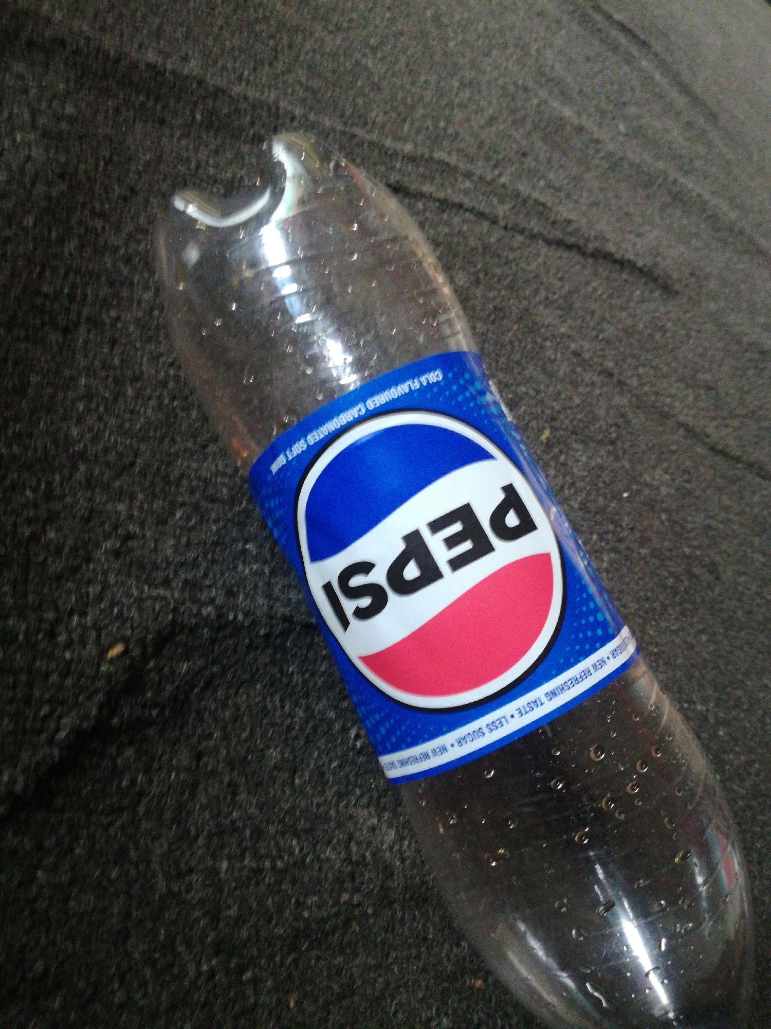 A clear plastic Pepsi bottle lies on a dark, textured surface, possibly carpet or asphalt, in a close-up, angled shot. The bottle appears mostly empty, with numerous small droplets of condensation or residual liquid clinging to its interior. The iconic Pepsi label is prominently displayed around the middle of the bottle. The label features the classic Pepsi logo: a red, white, and blue circular design with "PEPSI" written in white, sans-serif font across the white band.
Above the main logo, in blue text on a lighter blue background, are the words "COLA FLAVOURED CARBONATED SOFT DRINK". Below the main logo, along the bottom edge of the label, white text on a blue background reads "NEW REFRESHING TASTE • LESS SUGAR • NEW REFRESHING TASTE". The bottle itself shows slight indentations and wear consistent with a used plastic beverage container. The dark background is out of focus, emphasizing the bottle as the primary subject. There are no people or specific activities depicted; the scene is a still life of a discarded or finished beverage. The lighting suggests an indoor or shaded outdoor setting, but no specific time of day or location is identifiable.