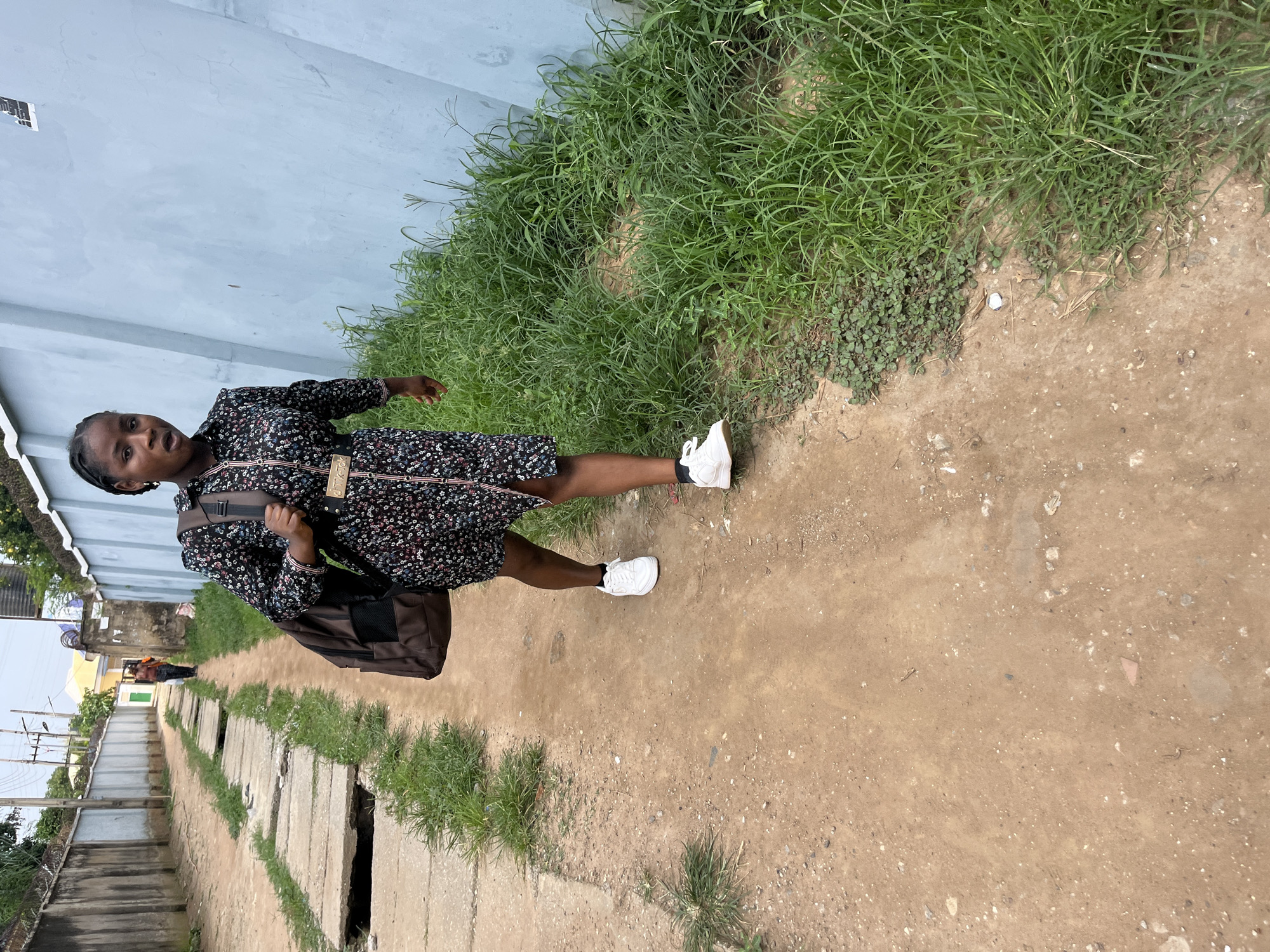 A young Black woman stands on a dirt path next to a wall and overgrown grass. She is wearing a dark, floral-patterned dress and white sneakers, with a dark brown messenger bag slung over her shoulder. The woman is looking at the camera with a neutral expression. In the background, the path continues, lined with concrete slabs that appear to cover a drainage system. Further down the path, other people and buildings are visible, suggesting an urban or semi-urban environment. The scene is outdoors, and the lighting indicates it is daytime, possibly overcast due to the lack of strong shadows. The presence of dense green grass and the overall atmosphere suggest a tropical or sub-tropical climate. The overall impression is of a candid moment captured while walking or pausing on a path.