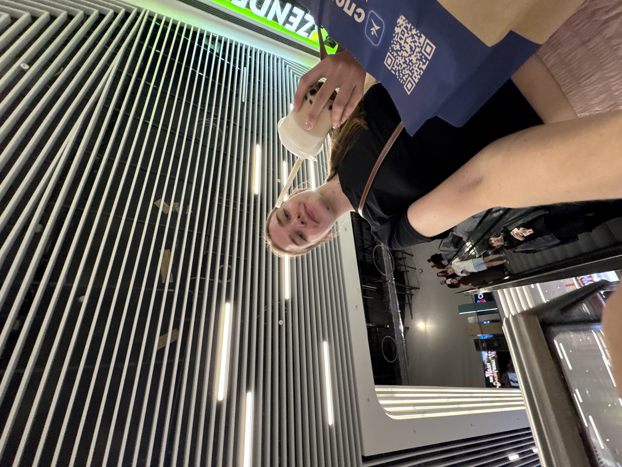 A young woman with blonde hair pulled back is taking a selfie on an escalator in what appears to be a modern shopping mall. She is wearing a black t-shirt and a pink skirt. She is holding a cup with a straw and a blue shopping bag. In the background, other shoppers are visible on escalators. The ceiling of the mall is covered with horizontal white slats. The location is identified as the city of Perm, Russia.