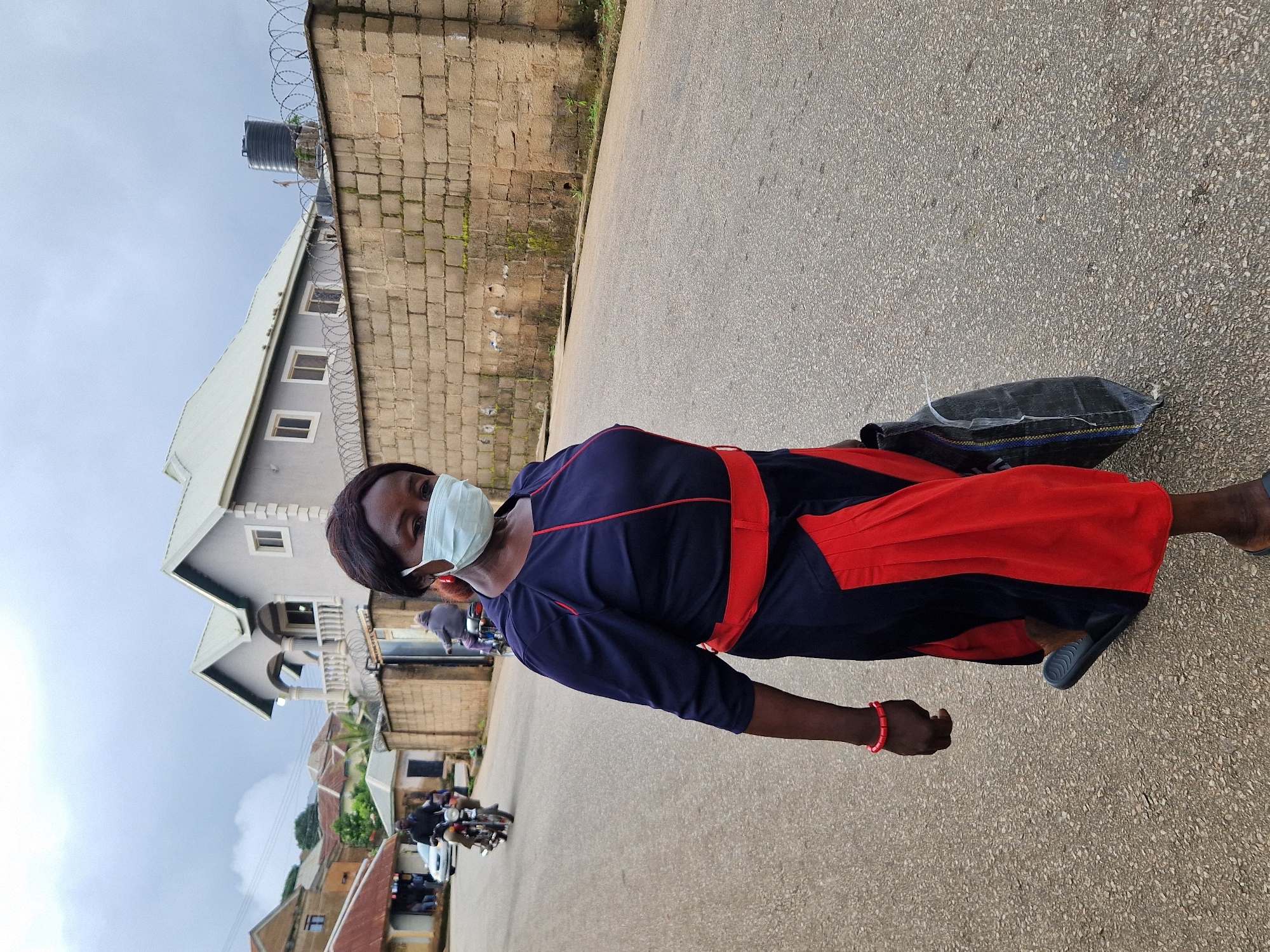 A woman wearing a dark blue dress with a red belt and red accents on the skirt, and a light blue face mask, walks on a paved street in Kabong, Nigeria. She carries a black bag in her right hand and wears a red bracelet on her left wrist. In the background, several residential buildings and a brick wall topped with barbed wire are visible. A water tank sits atop the wall. Two motorcycles with riders are also present on the street. The sky is overcast.