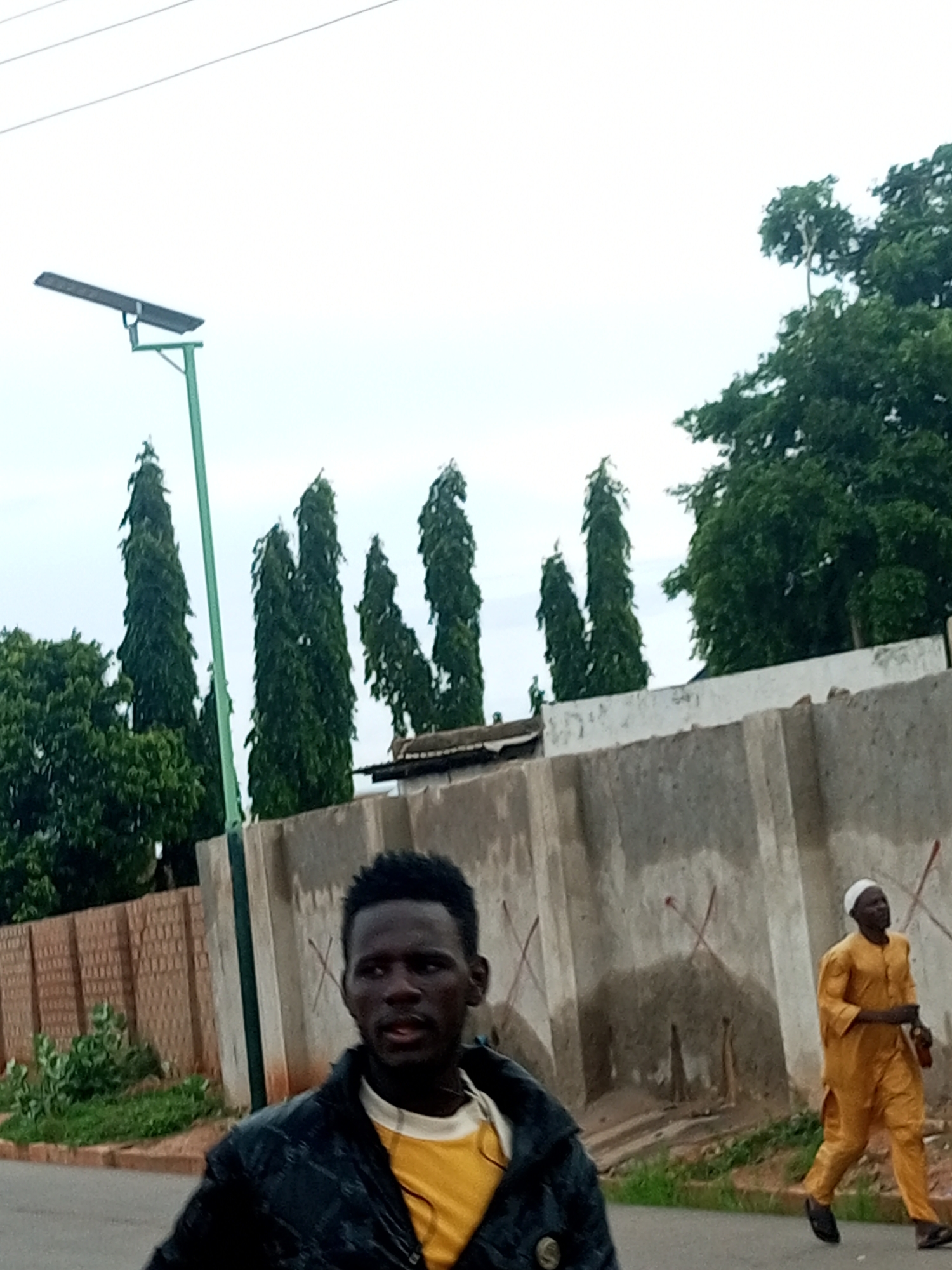 In Tumfure, Nigeria, a man wearing a black jacket over a yellow shirt is walking on a paved road, looking to his right. Behind him, another man in an orange traditional outfit and white cap is jogging past a concrete wall with red markings. Tall, dark green trees and a solar-powered street light are visible in the background, under an overcast sky. A brick wall borders the left side of the road.