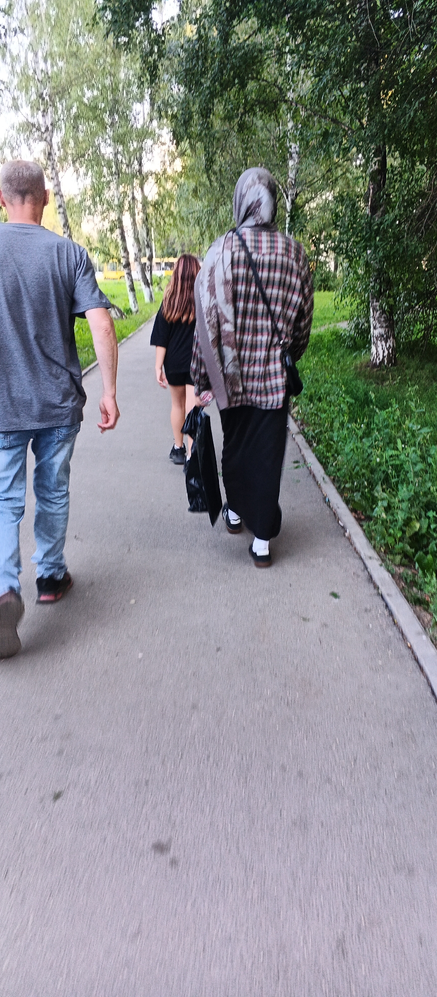 The image shows three people walking on a paved path in a park-like setting. The person on the left is a male wearing a grey t-shirt and blue jeans, with a black and red shoe. The person in the middle is a female with long, brown hair, wearing a black t-shirt dress and black shoes. The person on the right is wearing a headscarf and a plaid shirt over a black skirt, carrying a black bag. The path is bordered by green grass and trees. The scene appears to be in Izhevsk, Russia, given the context. The image was likely taken on a cloudy day, judging by the lighting.
