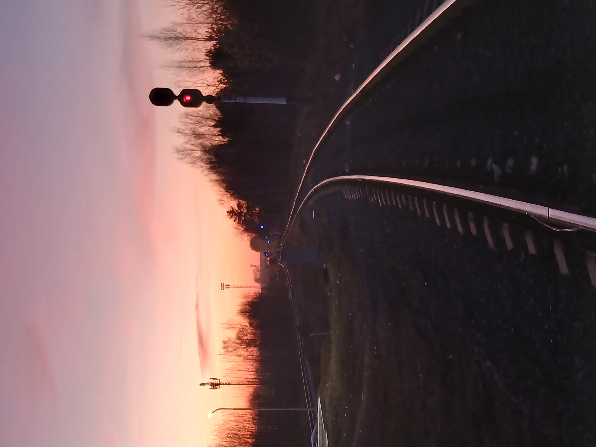 The image captures a railway track scene at twilight, either during sunrise or sunset. The perspective is looking down the tracks as they gently curve towards the right and recede into the distance.
On the left side of the tracks, a railway signal light is prominently featured, displaying a distinct red illumination, which typically signifies a stop or caution. Further along the track, more elements of railway infrastructure and signal posts are visible in the low light. The immediate ground beside the tracks, particularly on the right, is dark and appears to be earth or gravel.
A line of trees with bare branches forms the backdrop along the left side of the track, suggesting a cooler season such as late autumn or winter. The sky dominates the upper and left portions of the image, showcasing a beautiful gradient of colors that transitions from soft purples and pinks to warmer oranges and yellows near the horizon, characteristic of dawn or dusk.
In the far distance along the tracks, faint blue lights can be seen, possibly indicating another signal or a distant train. A street lamp is also visible towards the bottom-left of the frame. The overall ambiance is quiet and still, with no human figures or specific activities taking place. There is no visible text in the image.