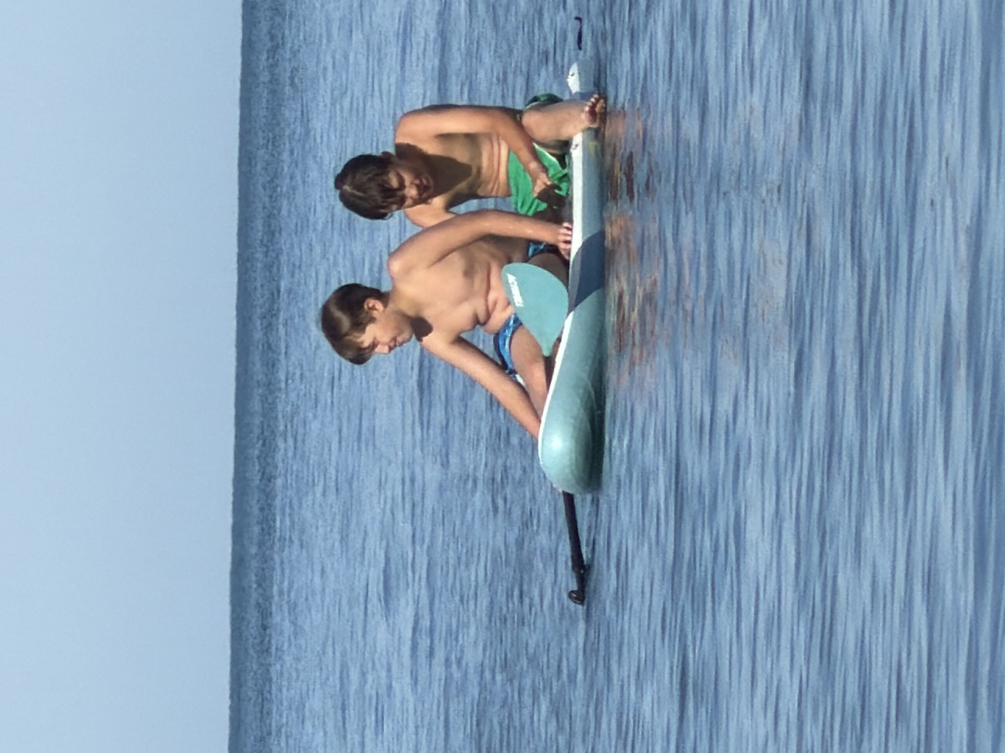 Two pre-adolescent males are seated on a light blue paddleboard in calm, blue water. The male on the left is wearing blue swim trunks and is holding a paddle. The male on the right is wearing green swim trunks. They are both looking to their left. The horizon line is visible in the background, where the water meets a light blue sky. The image does not provide any information to determine the location within the city of Partenit, Ukraine.