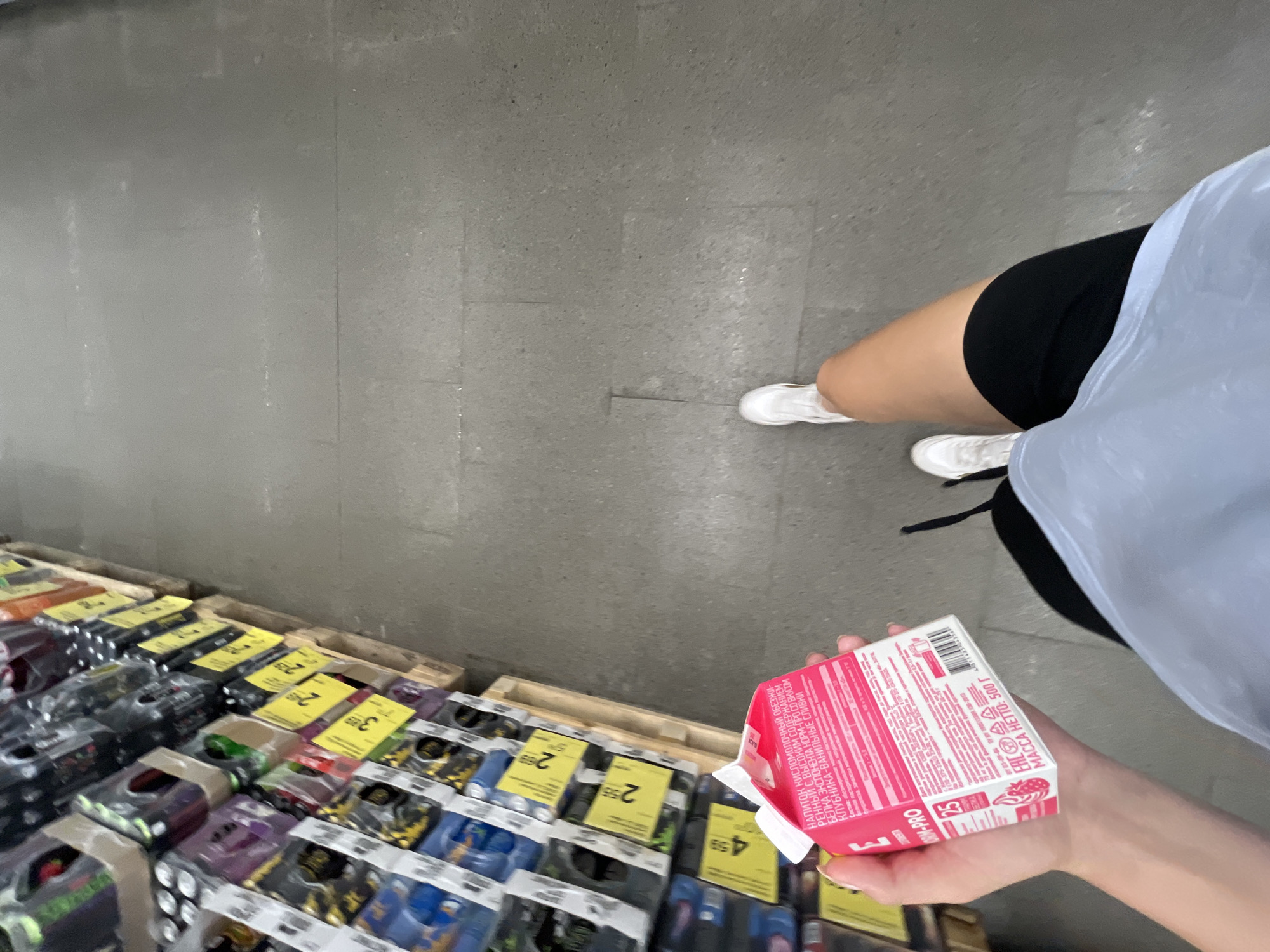 This first-person perspective media file captures a person's hand holding a small, pink carton of a fermented milk drink while standing in a supermarket aisle. The carton features Cyrillic text, with prominent mentions of "НАПИТОК КИСЛОМОЛОЧНЫЙ" (fermented milk drink), "БЕЛКА" (protein), and flavor details like "КЛУБНИКА-ВАНИЛЬНЫЕ СЛИВКИ" (strawberry-vanilla cream). The carton also displays nutritional information, including "МАССА НЕТТО: 500 Г" (net weight: 500g) and a barcode.
The background reveals rows of products on shelves, many of which are also packaged in plastic and have yellow price tags. One tag is clearly visible with the number "2" and "55". The flooring is concrete, suggesting an indoor retail environment. The person is wearing white sneakers and black shorts, and a pale blue garment is visible. The lighting appears to be standard for a supermarket, indicating it could be daytime or evening. The overall scene is mundane, focusing on a shopping activity within a store in Babruysk, Belarus, as suggested by the location context.