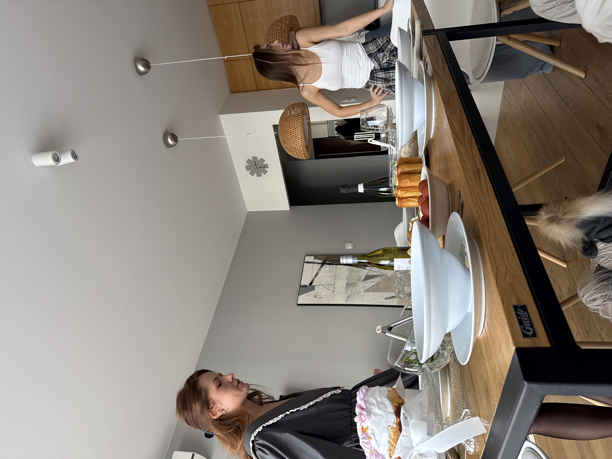 The image shows an indoor scene in what appears to be a modern apartment, possibly located in Warsaw, Poland, based on the visible interior design style, though specific local landmarks are not visible.
Two women are present. One is wearing a white tank top and plaid pants and appears to be reaching across a table. The other is wearing a dark dress with white trim and is standing on the opposite side of the table.
The central focus is a long wooden table set for a meal, with white plates, bowls, cutlery, and glassware. Food items, including what appears to be pastry and fruit, are arranged along the table. Bottles of wine are visible. A piece of framed art is hung on the wall in the background. Two pendant lights with woven shades hang from the ceiling, as well as several other modern light fixtures. A white chair is adjacent to the table.