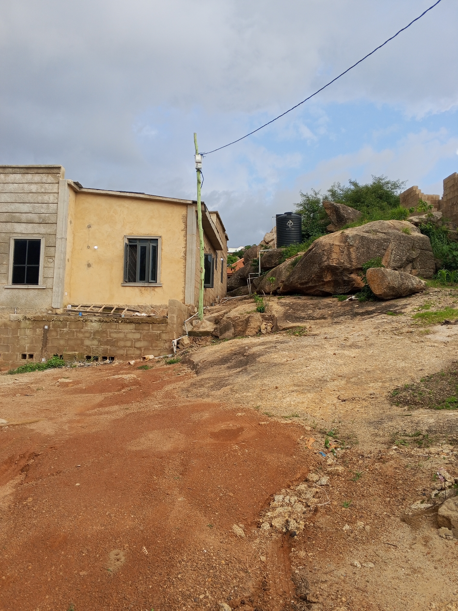 The image captures an outdoor scene in Kabong, Nigeria, featuring a partially constructed building and a rocky, uneven terrain. The building on the left appears to be a single-story structure with a yellow-painted facade and a section with textured concrete walls. It has dark-framed windows. Adjacent to it, to the right, is another section of the building, also under construction, with visible brickwork.
The foreground is dominated by a reddish-brown, dusty ground, likely soil or gravel, with some scattered debris. The middle ground is characterized by a large, weathered rock formation with various sizes of boulders and smaller stones, interspersed with green vegetation. A tall, black water tank is positioned on top of the rocks. A green pole with electrical equipment is visible near the building, and a blue wire stretches across the upper part of the frame. The background consists of a cloudy sky with patches of blue. The overall impression is of an undeveloped or developing area.