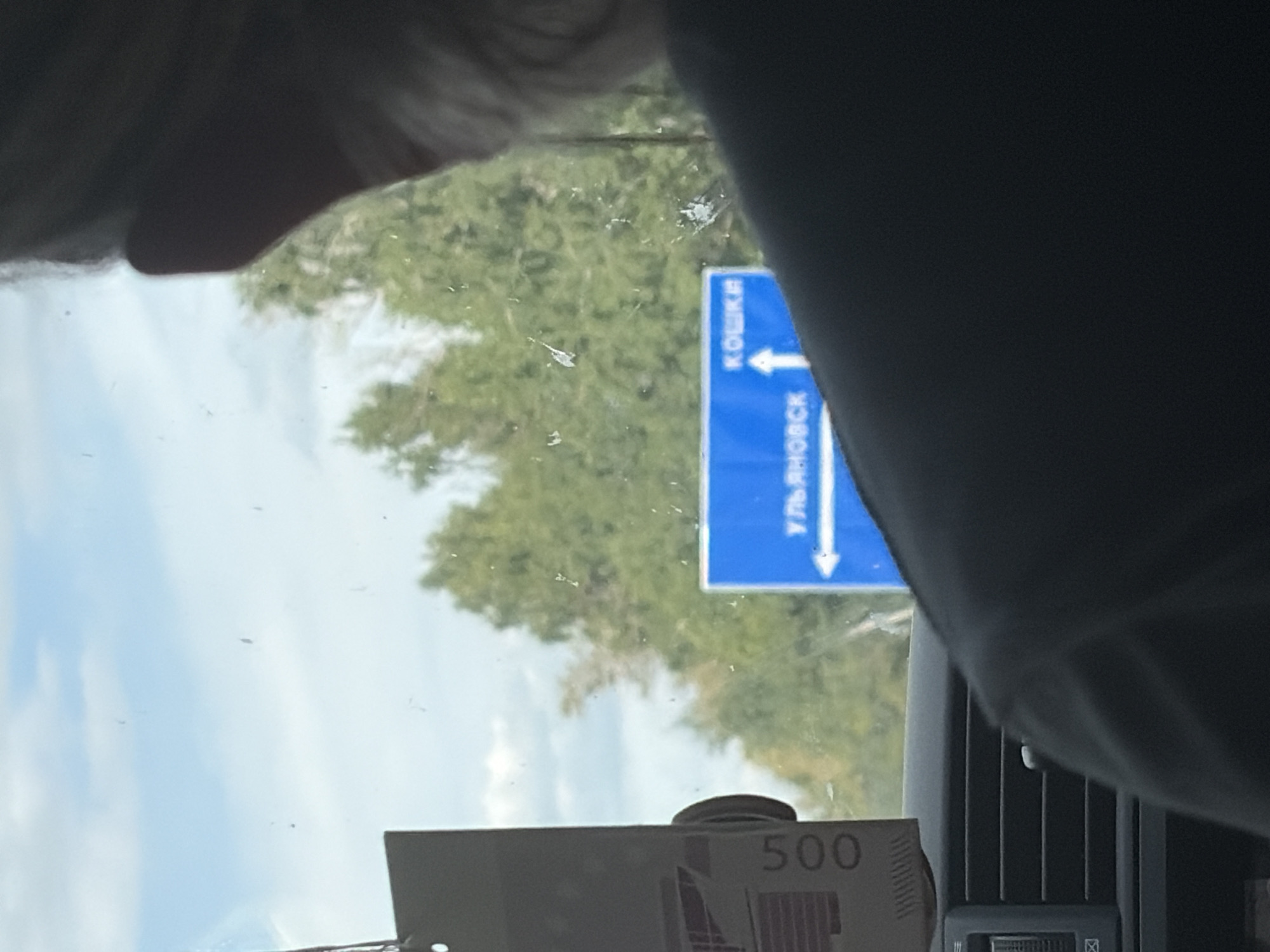 The image shows a view from inside a vehicle, looking out through the windshield. A person's shoulder and the back of their head are visible on the right side of the frame. A blue road sign is visible through the windshield, indicating directions to "КОШКИ" (Koshki) and "УЛЬЯНОВСК" (Ulyanovsk). The sign has an upward arrow pointing towards Koshki and a leftward arrow pointing towards Ulyanovsk. A tall, dark object with the number "500" printed vertically in white is attached to the windshield on the left side. Trees and a cloudy sky are visible in the background. The image appears to be taken in Russia, potentially near the city of Vershiny, as suggested by the presence of Russian text on the road sign.