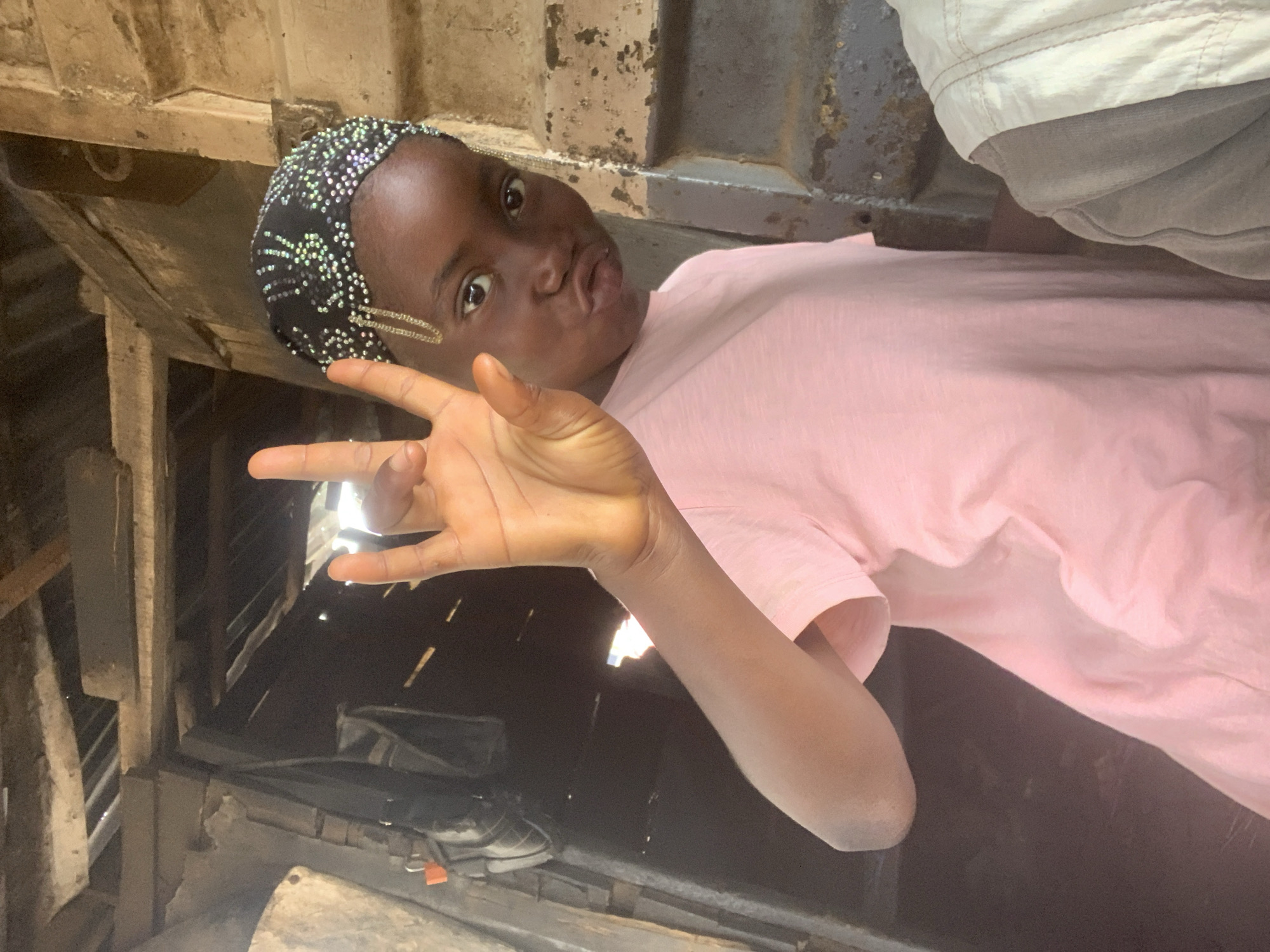 The media file is a portrait of a young Black individual, likely female, wearing a pink t-shirt and a black headscarf adorned with small, shiny beads. The person is positioned in a slightly reclined, informal pose, making a "peace" sign with their right hand. Their lips are pursed, suggesting a playful or coquettish expression. A golden hair clip is visible on the left side of their forehead, adding a subtle decorative detail. The background is rustic and appears to be an outdoor or semi-outdoor setting. There are wooden structures and possibly corrugated metal elements, indicating a possibly improvised or informal shelter. Beneath the subject, what appear to be shoes are visible in a dark, cluttered area. The lighting suggests it might be daytime, with some natural light filtering into the scene. The overall impression is of a casual, candid moment captured in what seems to be a less developed environment. The location context of Shagamu, Nigeria, suggests a West African setting.