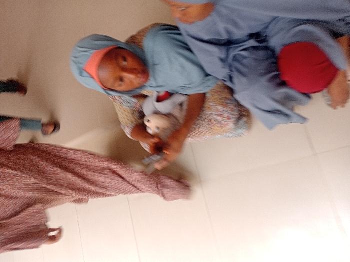 The scene depicts an indoor environment with a light-colored tiled floor in the city of Kwazailewa, Nigeria. A young female, appearing to be a child or teenager, is seated in the central foreground, wearing a patterned garment, a light blue hijab, and an orange head covering. She is observed holding two objects: a small, light-colored toy resembling a doll with a brown head and grey body in one hand, and a dark, rectangular object, possibly a mobile phone, in her other hand. Her gaze is directed downwards towards these items. To her immediate right, another individual is partially visible, seated, clad in a blue garment with a red item positioned on their lap; only their bare feet are discernible in the lower right. In the background, two additional individuals are in motion. The lower body of one person, dressed in a long, pleated, reddish-brown garment, walks from left to right. Further back, the feet, wearing dark sandals, and lower legs, clad in light blue trousers, of a fourth person are partially visible.