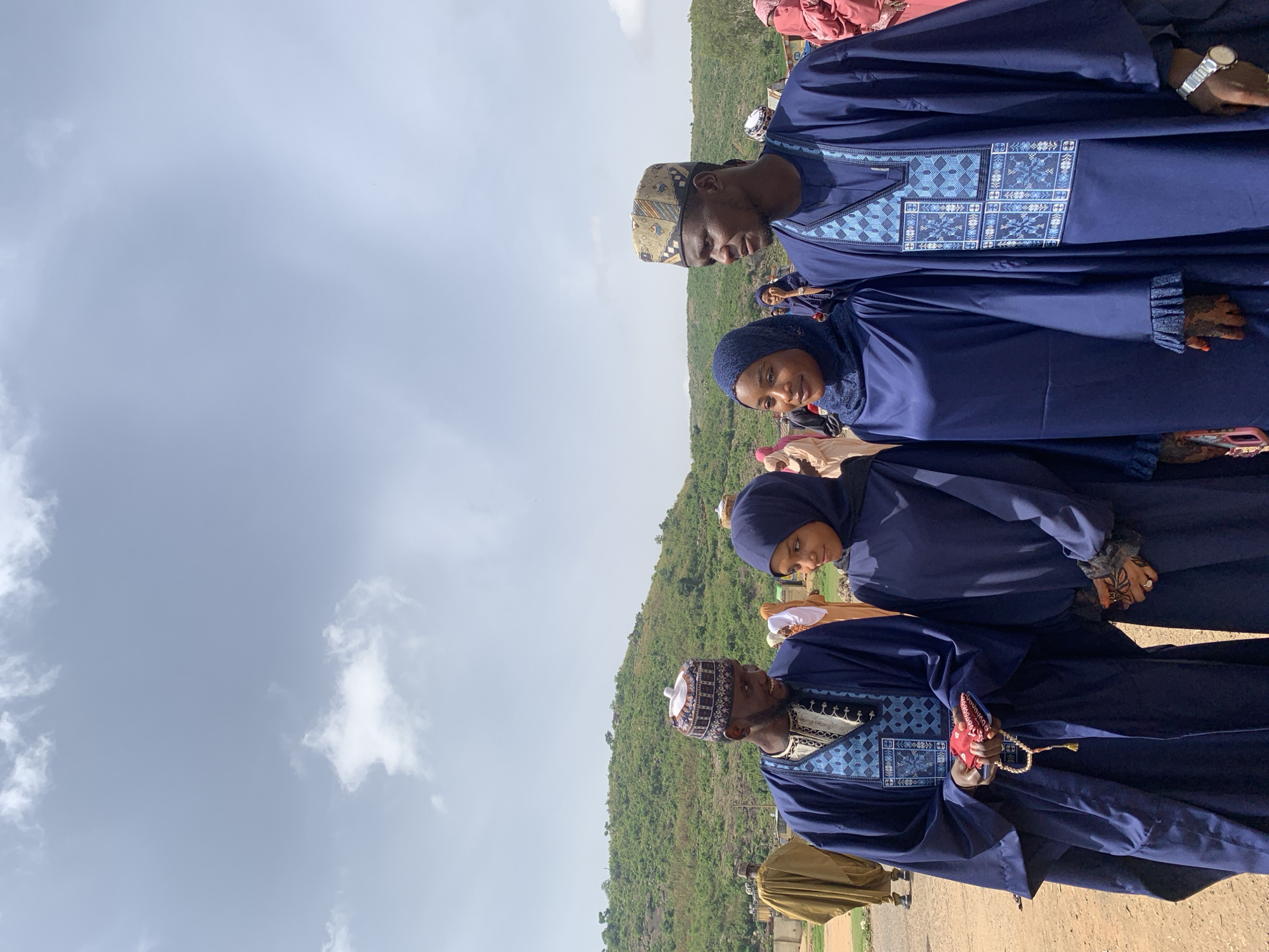 The image shows a group of people in Naraguta Hausawa, Nigeria. Four people are in focus, all wearing blue garments. The two men are in traditional attire with detailed embroidery. The two women wear hijabs. The background shows a partly cloudy sky and a green hillside. The scene is captured outdoors, likely during the day.