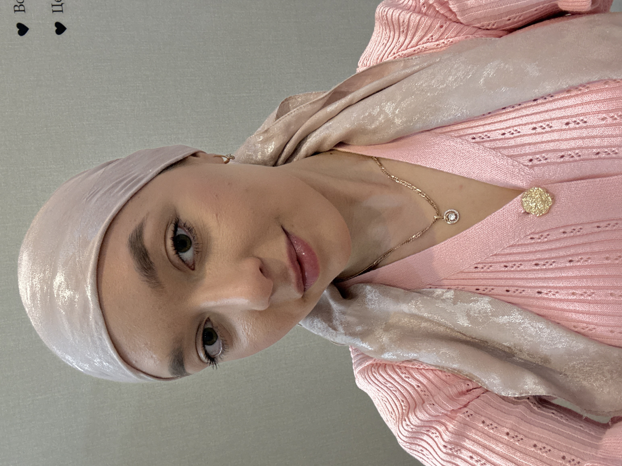 A woman is centered in the image wearing a pink headscarf, a gold necklace with a diamond pendant, and a pink knit sweater with a gold floral brooch. The background is a solid light gray color. The location is not discernible as Урмаево, Russia.