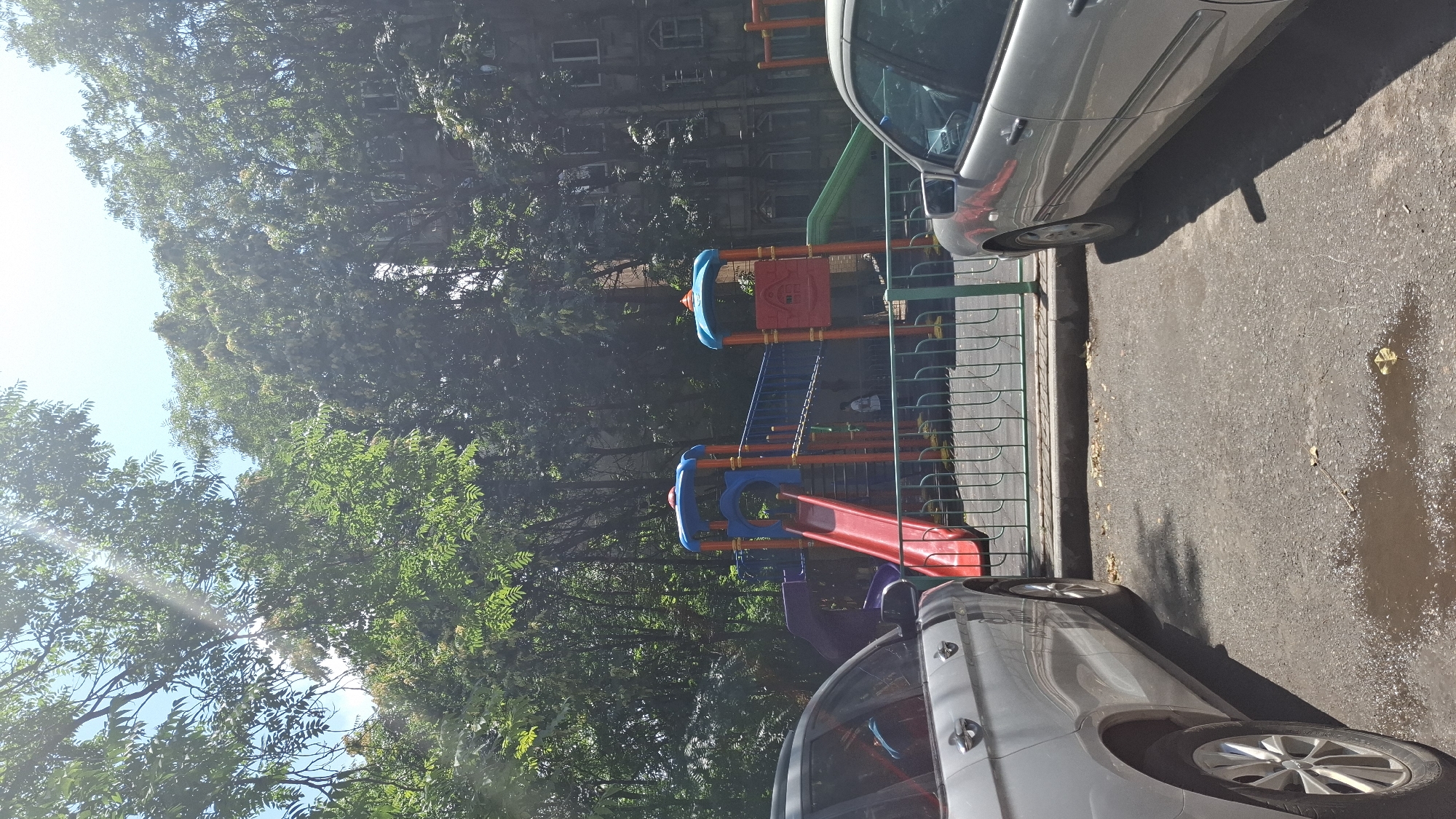 The image captures a sunny day in an urban environment, possibly in Shymkent, Kazakhstan. In the foreground, two silver cars are parked on an asphalt surface. One car is a larger SUV on the left, and a sedan is on the right. The ground shows some wet patches, suggesting recent rain or cleaning. Behind the cars, a brightly colored children's playground is visible, featuring slides, a climbing structure, and a bridge. The playground is enclosed by a green metal fence. Lush green trees surround the playground and extend upwards, partially obscuring the facades of apartment buildings in the background. The sunlight streams through the tree canopy, creating dappled light on the scene. A few people, appearing as small figures, are present on or near the playground. The overall impression is one of a residential area with recreational facilities for children.