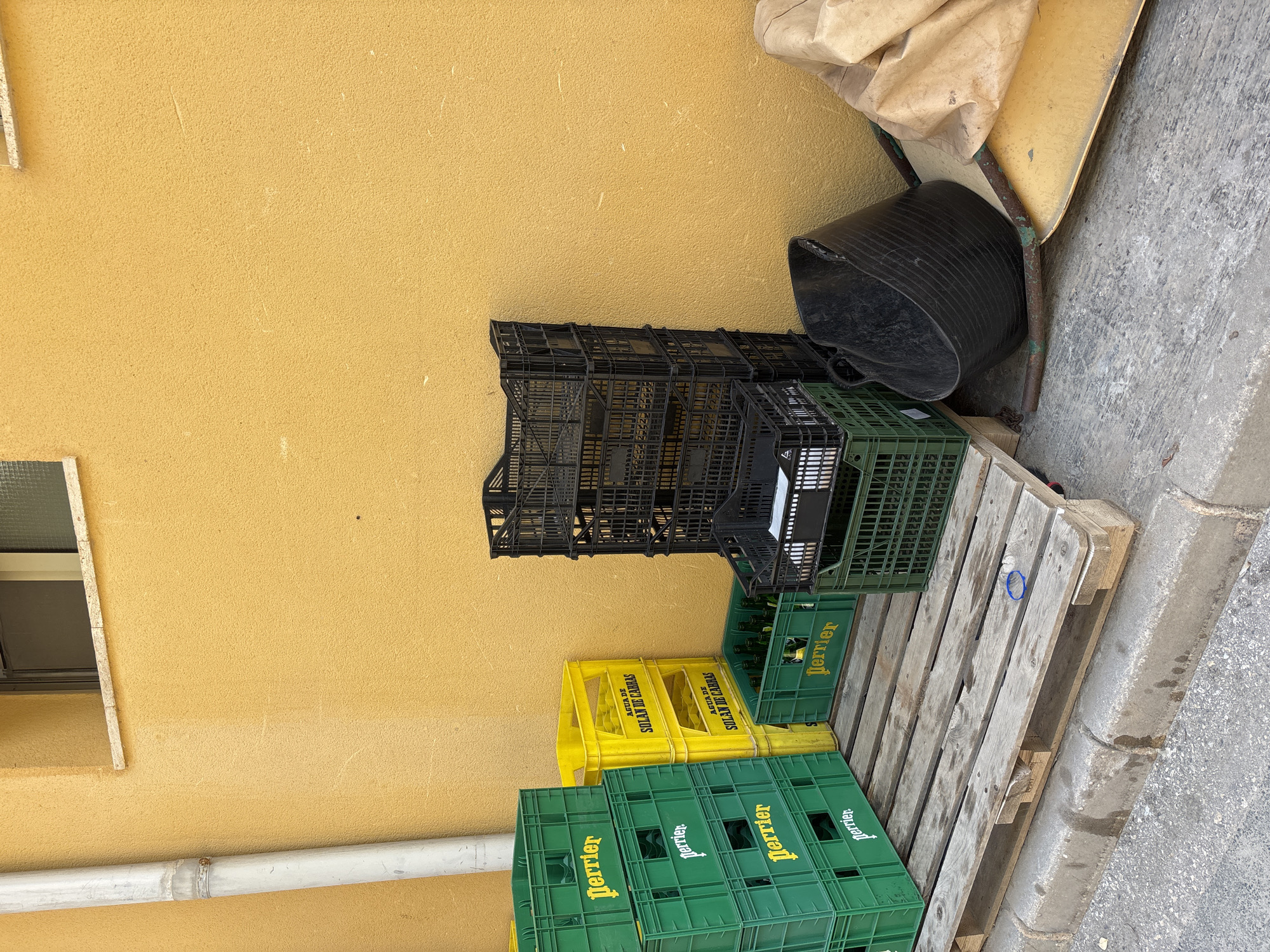 In an outdoor urban setting in Orihuela, Spain, several stacks of plastic crates are arranged against a textured yellow wall. To the left, a series of green Perrier crates are piled high. Adjacent to these, a stack of yellow crates, labeled "AGUA DE SOLAN DE CABRAS," is placed on a wooden pallet. In the center, a tall stack of black plastic crates dominates the mid-ground, with a smaller green crate containing bottles on a pallet to the right. To the right of the crates, a black plastic tub and a large, light brown tarp are visible on the ground. The scene is captured in daylight, with a concrete sidewalk and curb in the foreground, and a downspout running vertically along the left edge of the wall. The overall impression is one of storage or discarded items.