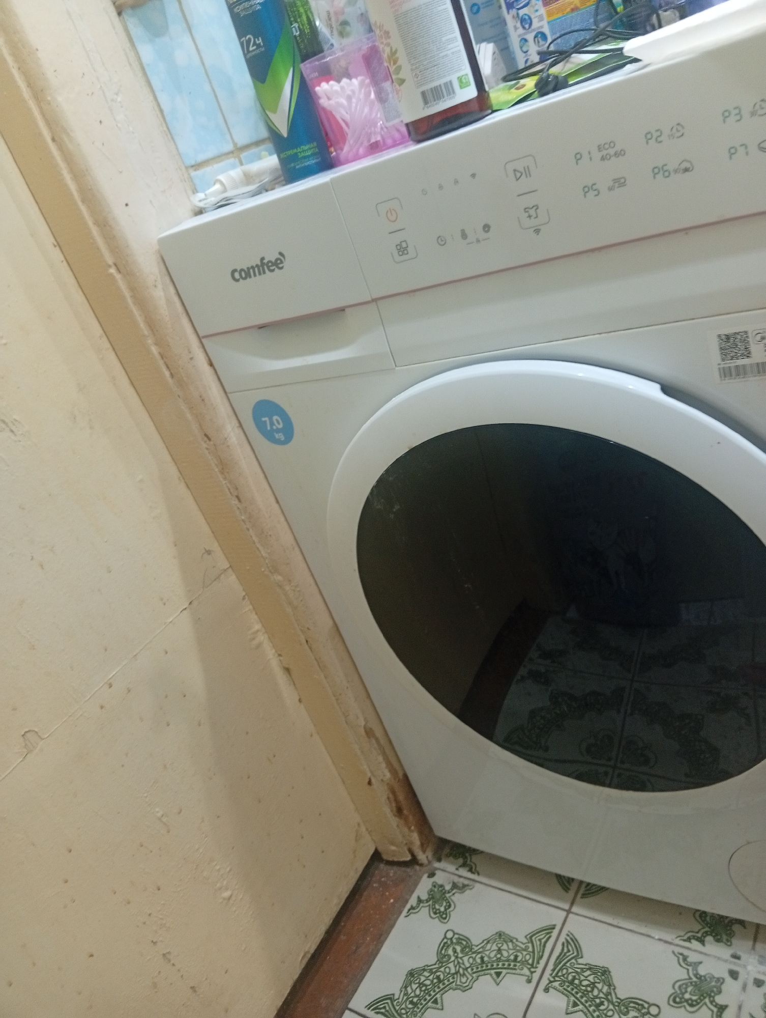 A white Comfee washing machine is positioned in a room. The washing machine has a control panel with various program selections labeled P1 through P7 and an "ECO 40-60" option. A circular sticker with "7.0 kg" is visible on the front of the machine. The room has tiled flooring with a green decorative pattern. The wall behind the washing machine is tiled with blue and white squares. To the left of the washing machine, a textured, light-colored wall is visible, possibly plaster. Several toiletry items, including a blue can with "72H" and a pink container with cotton swabs, are placed on top of the washing machine. The overall scene suggests a domestic environment, likely a bathroom or laundry area. The specific location of Pushkino, Russia, is an external detail not deducible from the image itself.