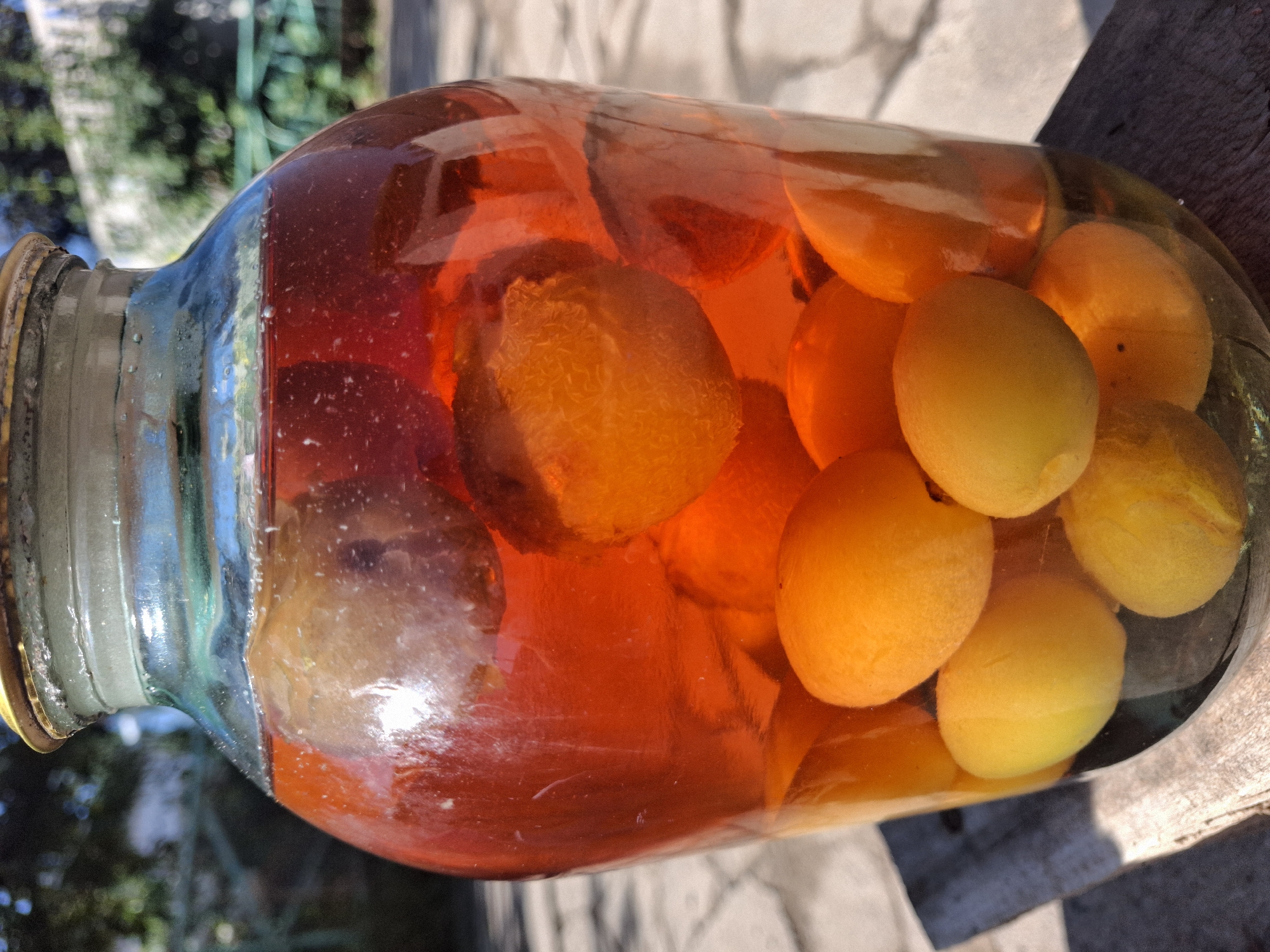 A glass jar filled with a reddish-orange liquid and whole apricots is positioned outdoors on a wooden surface. The jar is sealed with a metallic lid. The background shows blurred foliage and a building, suggesting an outdoor, possibly rural or suburban setting. The lighting indicates it is daytime. The location in Jeti-Ögüz, Kyrgyzstan, is not visually discernible from the image itself.