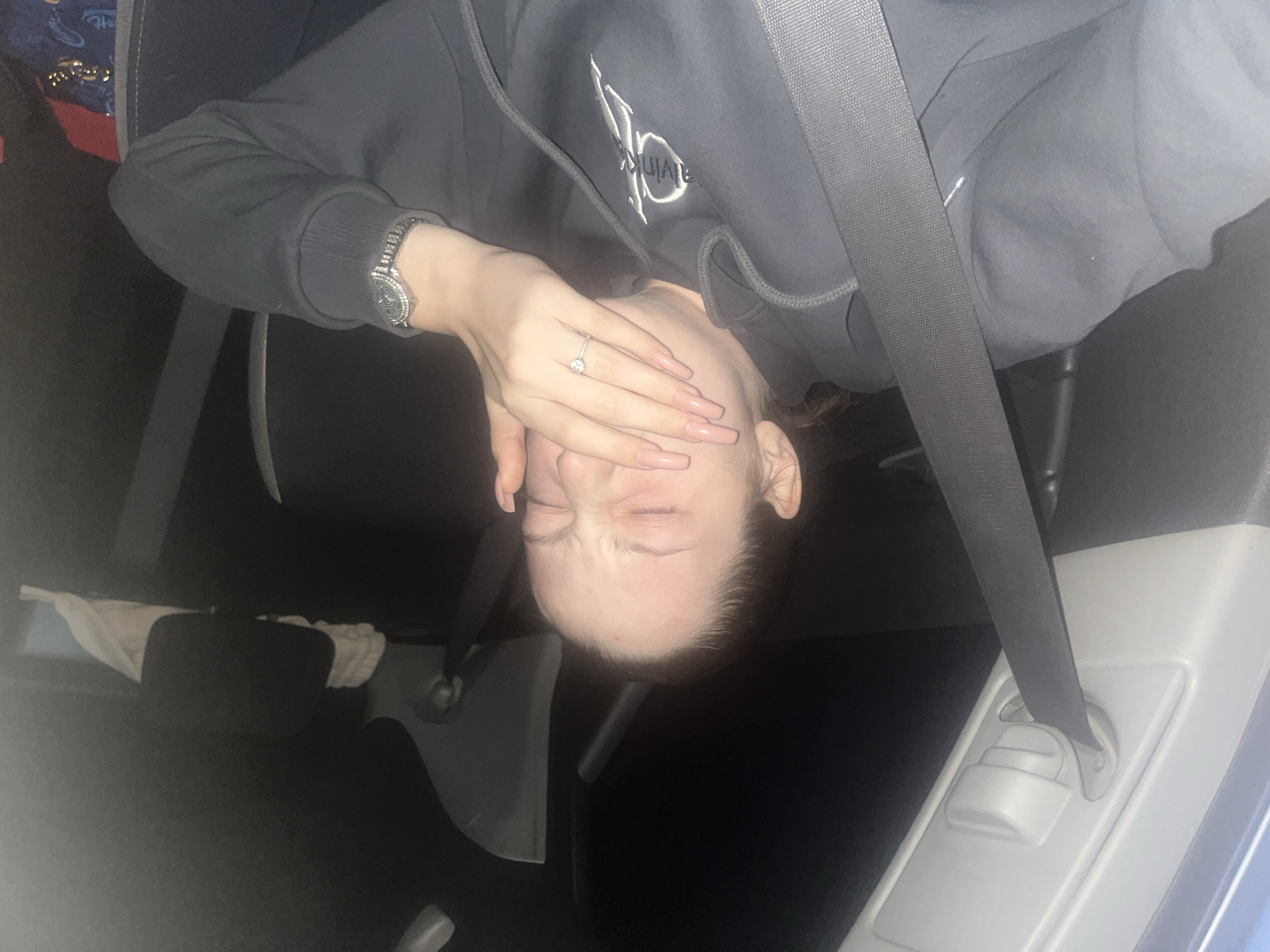 The image displays a person from the chest up, possibly lying down or captured in a rotated perspective, inside what appears to be a car interior. The person wears a dark grey hooded sweatshirt featuring a white, stylized logo on the chest, resembling "CK" or "Calvin Klein."
Their right hand covers the lower part of their face, with fingers slightly splayed. This hand shows long, light pink fingernails and a ring with a single clear stone on the ring finger. A watch with a shiny, possibly jeweled, band and face is visible on their left wrist. The person's eyes are closed, and their forehead is wrinkled, suggesting an expression of grimace, exasperation, or discomfort.
A grey seatbelt stretches across their torso, indicating they are in a vehicle. The background reveals dark fabric car seats, a dark headrest with a white cloth draped over it, and a light-colored plastic panel where the seatbelt is anchored. A bag with red and gold patterns is partially visible in the upper left background. The overall lighting is dim, suggesting it is nighttime or a low-light environment within the car. No specific text other than the stylized logo is clearly legible.