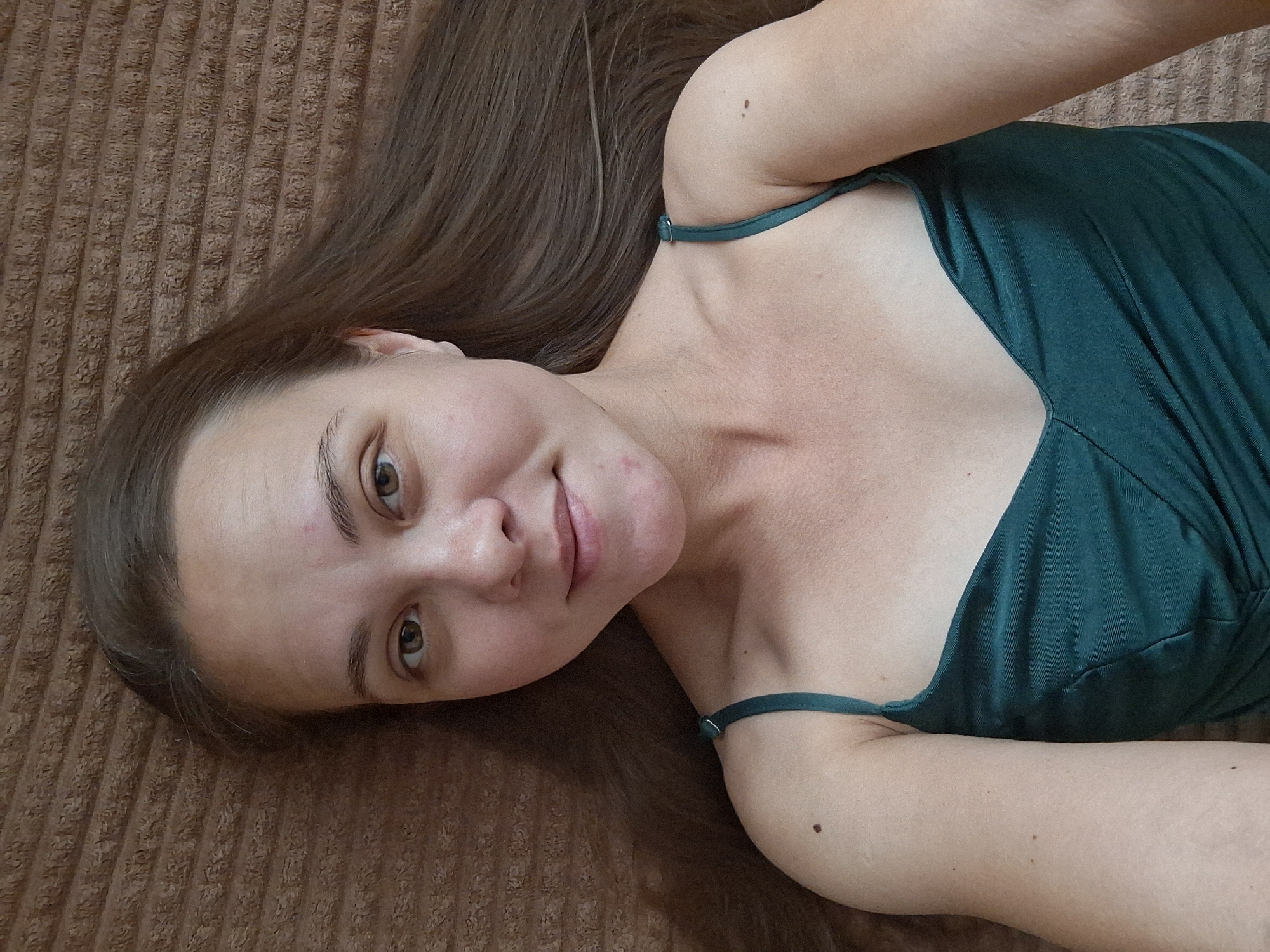 A young woman with long brown hair is lying on a brown, corduroy-like surface, looking up at the camera. She is wearing a dark teal, strappy dress. Her head is tilted, and she has a gentle smile. Her eyes are brown, and her eyebrows are well-defined. The lighting suggests an indoor setting, possibly during the day. There is no visible text in the image. The overall mood is relaxed and intimate.